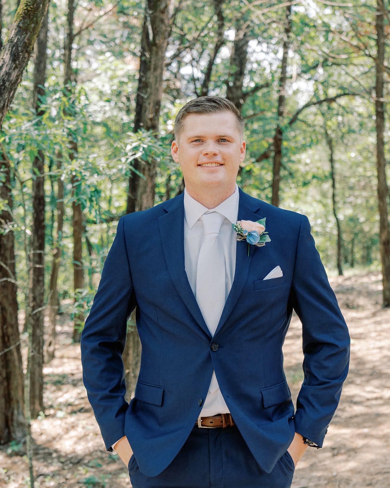 A person wearing a navy blue suit and white shirt poses in a wooded outdoor setting for a formal portrait.