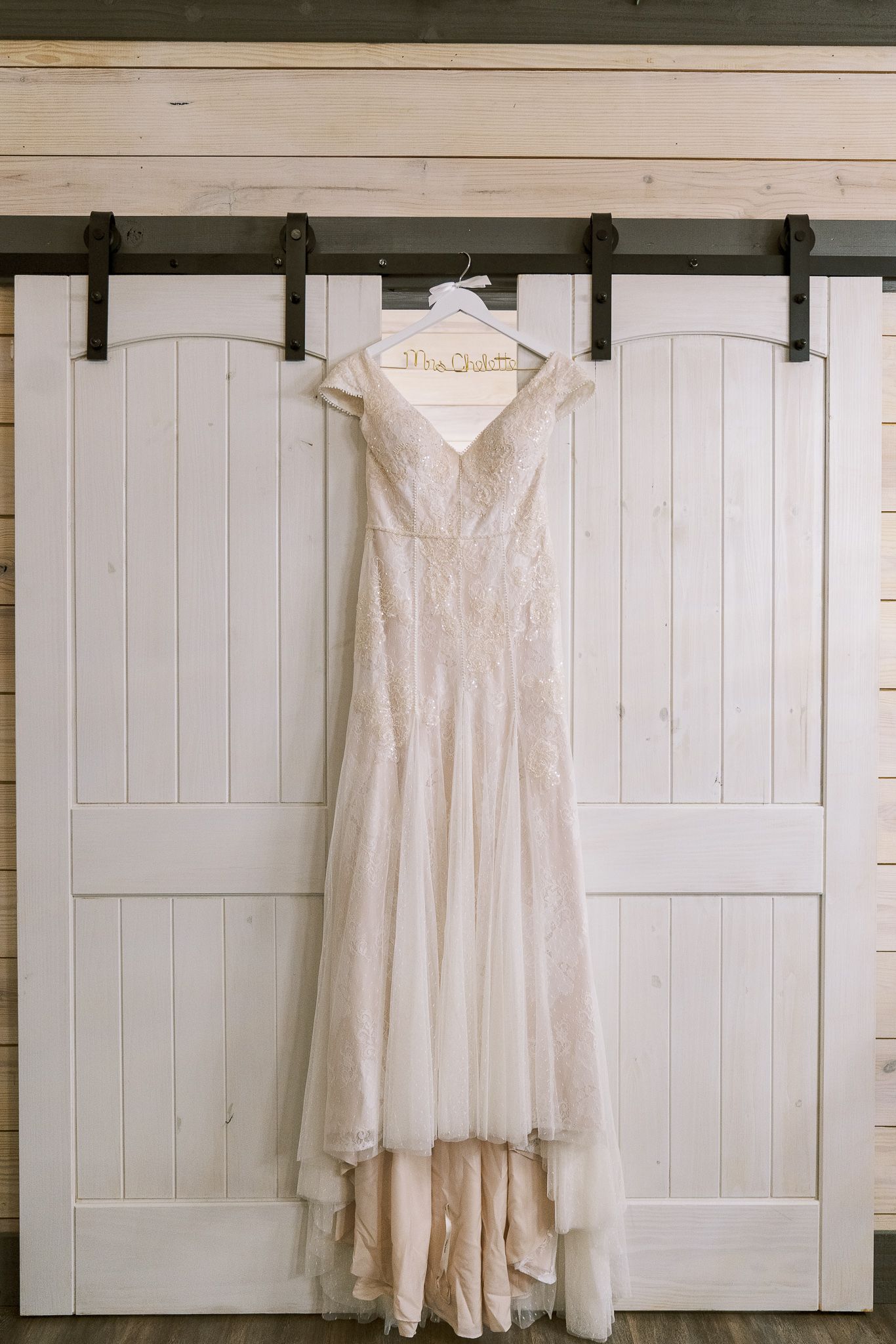An elegant white wedding dress with lace details hangs on a rustic barn door with sliding hardware.
