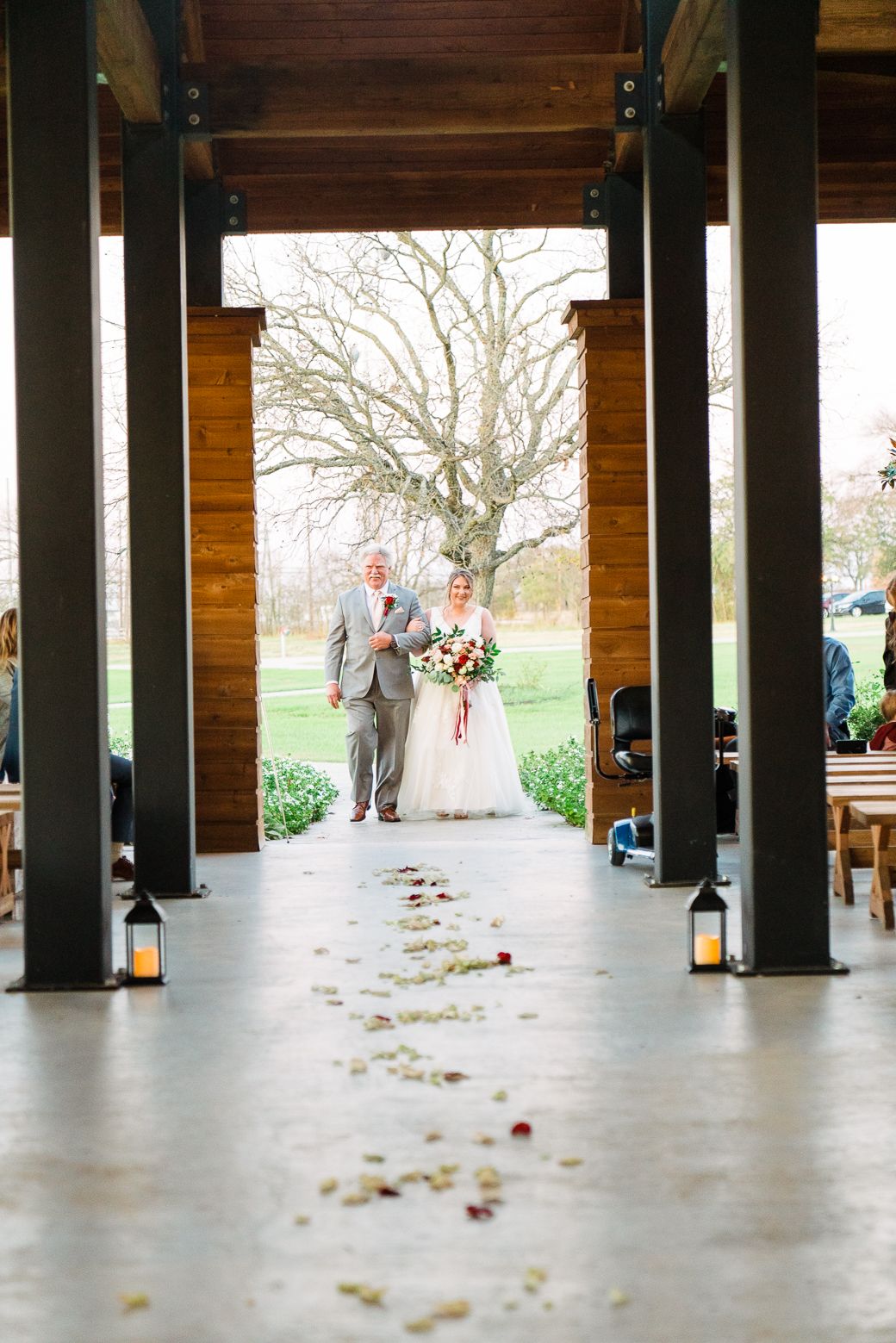 A wedding ceremony aisle lined with lanterns and flower petals leads to a rustic wooden pavilion framing a bare tree.