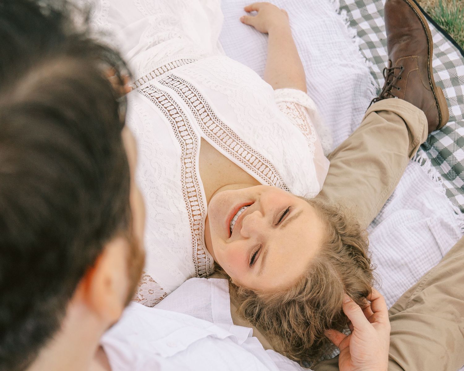 Woman in white lace dress laughing while lying on a blanket outdoors during an engagement photo session.