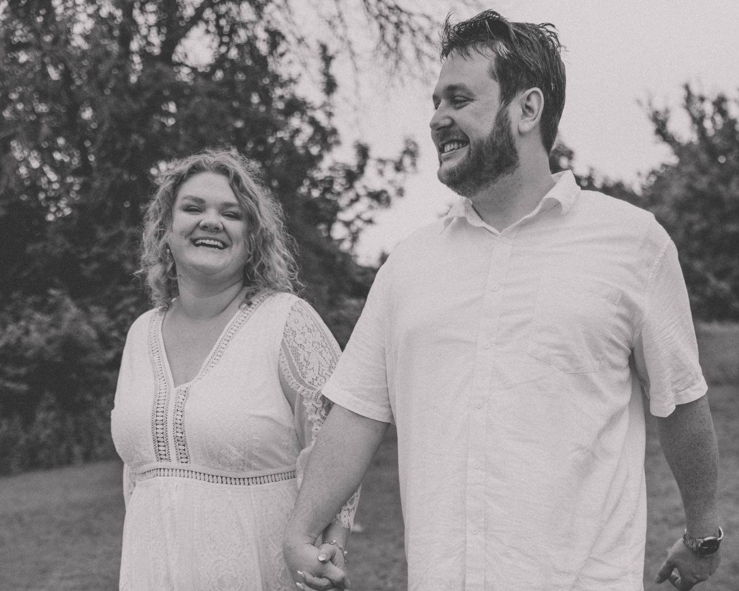 Black and white photo of laughing couple holding hands and walking together through a lush green park.