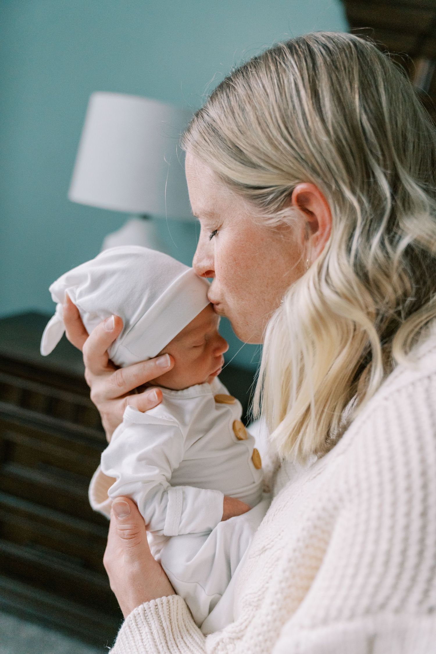 A parent in a white sweater holds and kisses a newborn baby wrapped in white blankets against a teal wall background.