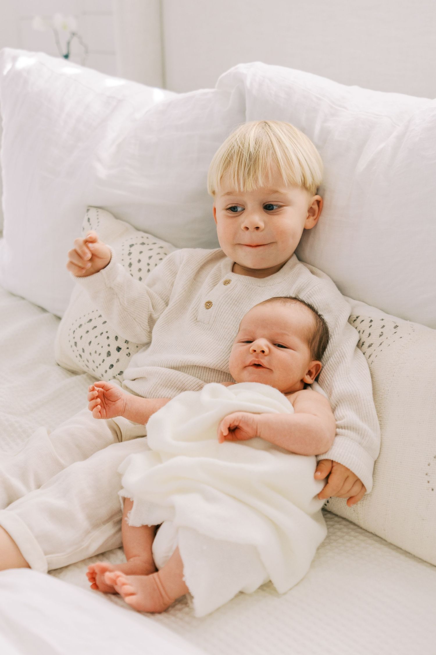 Two young children in white outfits rest together on a plush white couch.