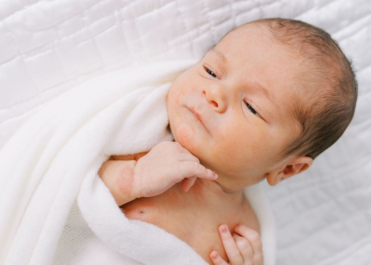 Sleeping newborn baby wrapped in soft white blanket rests peacefully on white bedding.