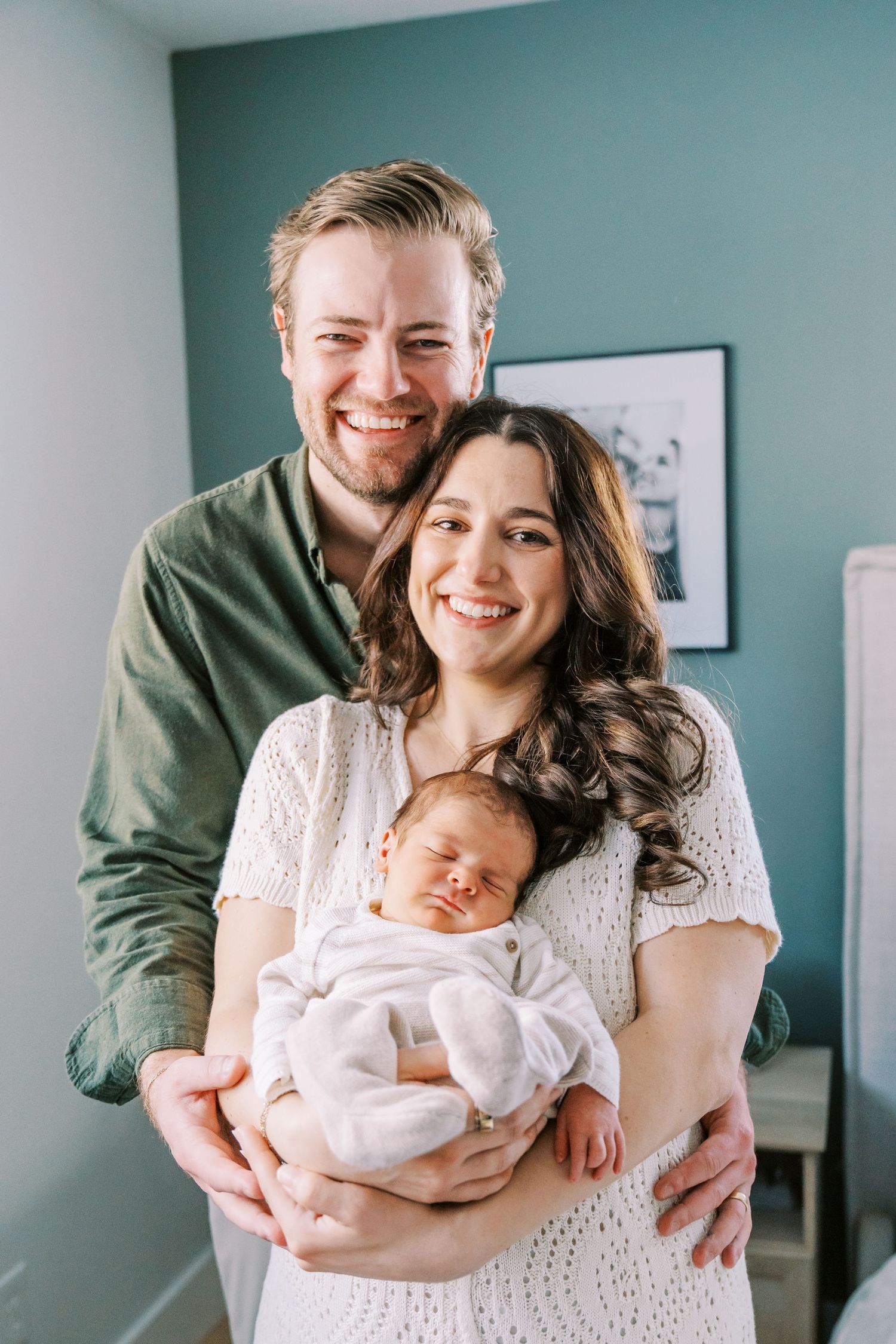 A couple stands together holding their sleeping newborn baby in a room with teal walls and framed artwork.