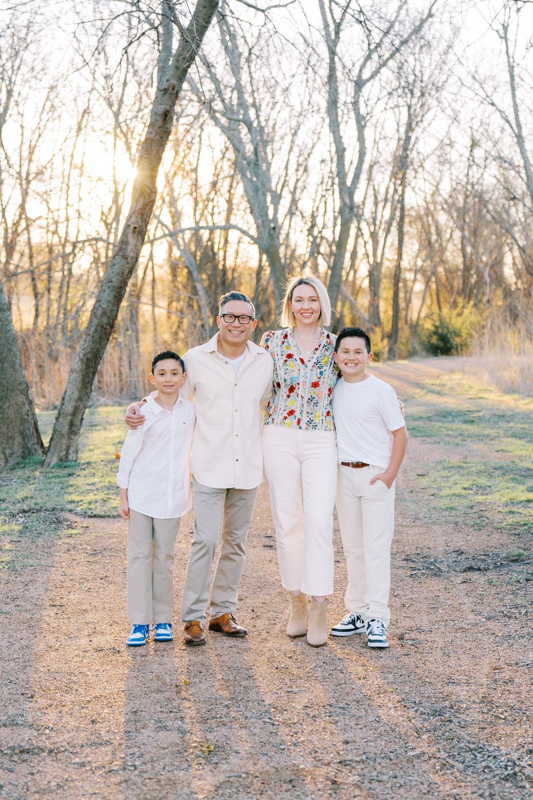 A family of four poses together on a dirt path surrounded by bare trees at sunset in coordinated light-colored outfits.