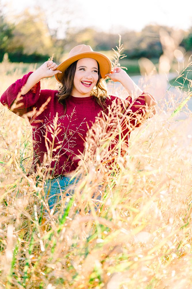 Someone wearing a burgundy sweater and straw hat lies in golden autumn grass during a sunny outdoor photoshoot.