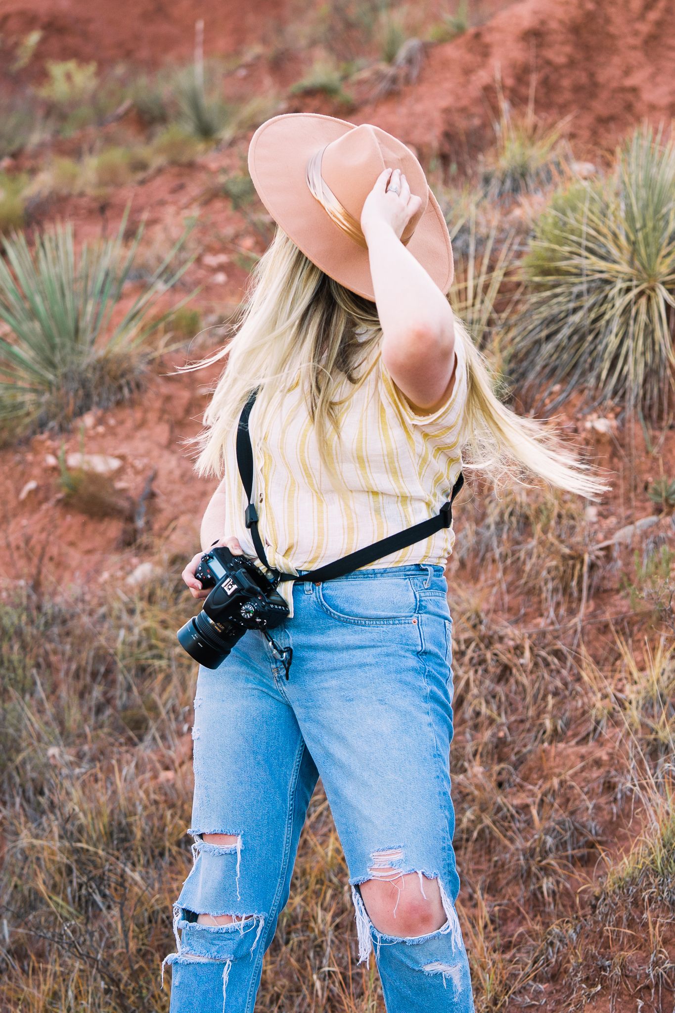 Person wearing ripped blue jeans and striped top carries camera in desert landscape with red rocks and desert plants.