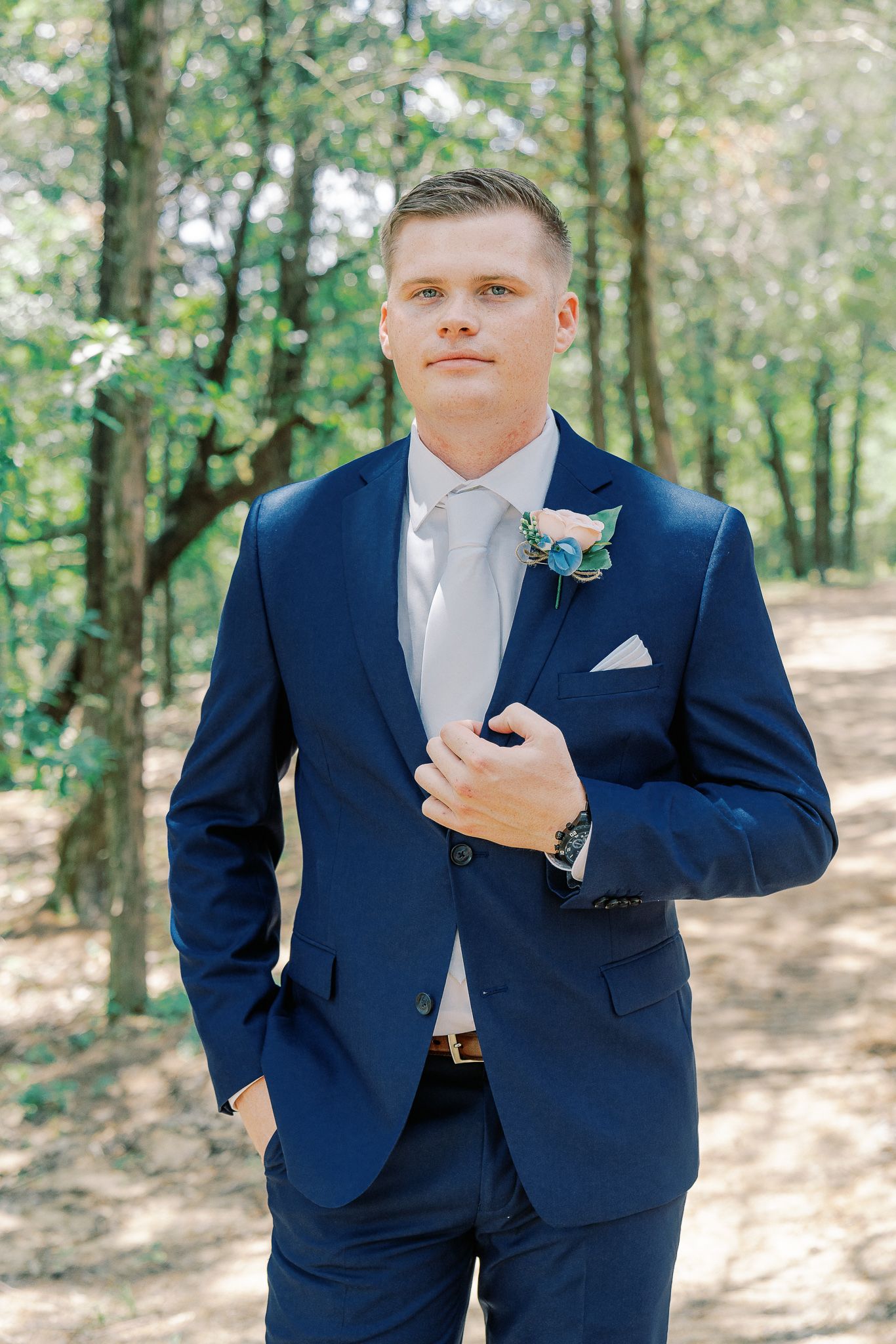 A groom in a navy blue suit and light gray tie stands in a wooded outdoor setting for wedding portraits.