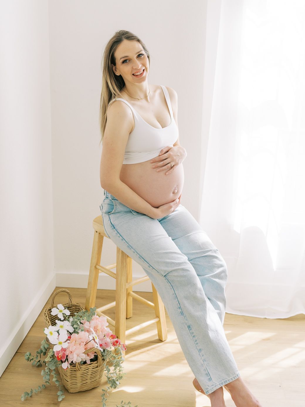 Maternity photoshoot featuring floral basket props and natural window lighting with casual denim styling.