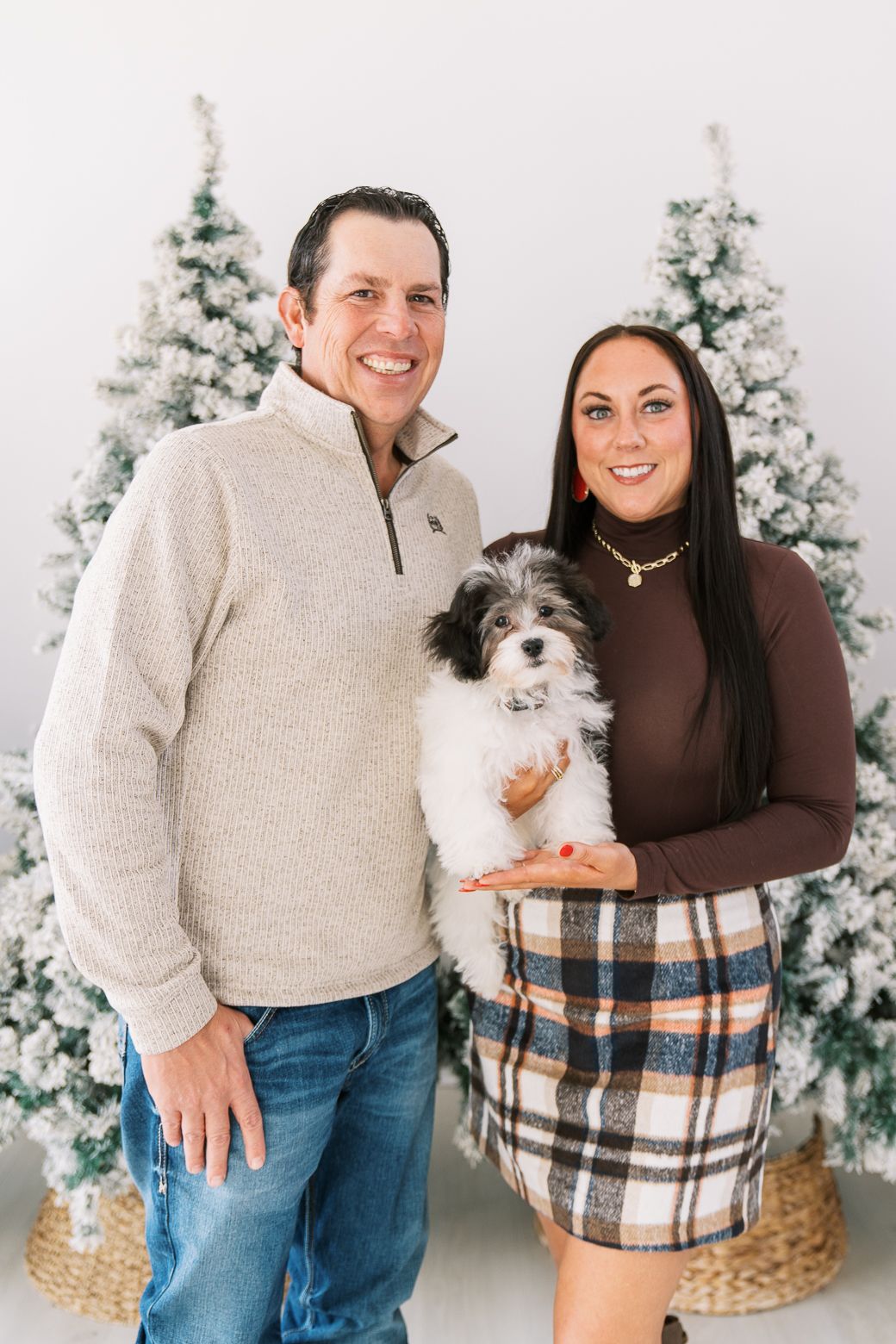 A couple holds their small black and white dog while standing in front of flocked Christmas trees for a holiday portrait.