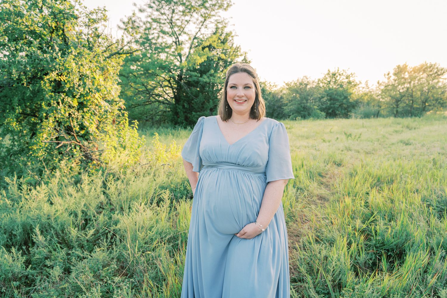 Expectant mother in flowing powder blue dress poses gracefully in a sunlit meadow during golden hour.