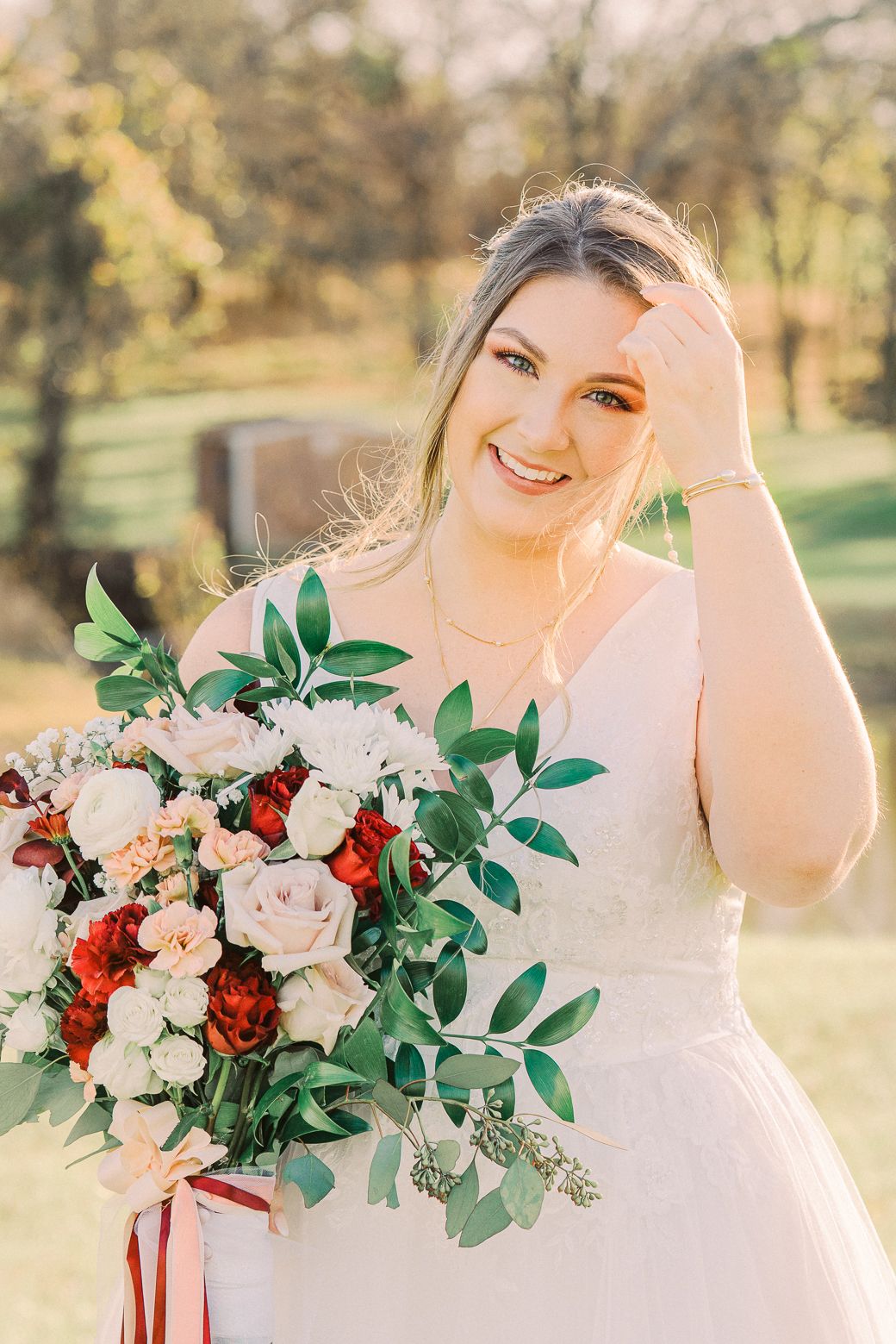 Bride poses with elegant red and white bouquet featuring roses and eucalyptus in outdoor garden setting.