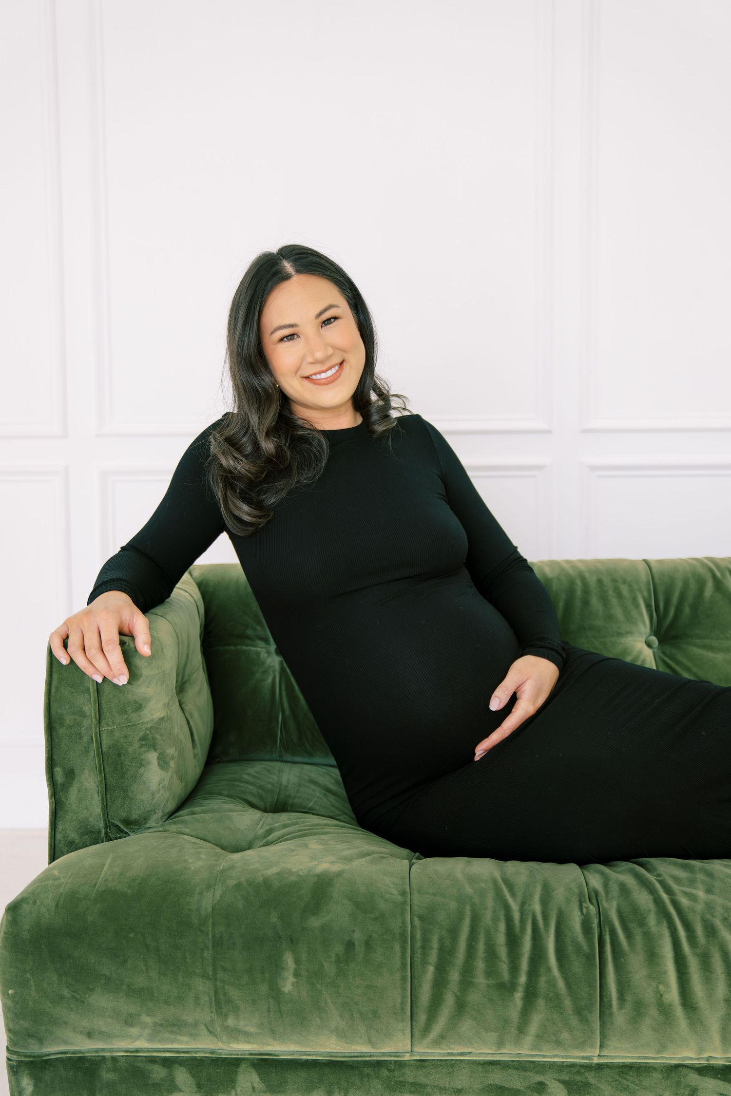 Smiling pregnant woman in black dress resting on green velvet sofa during maternity photo session.