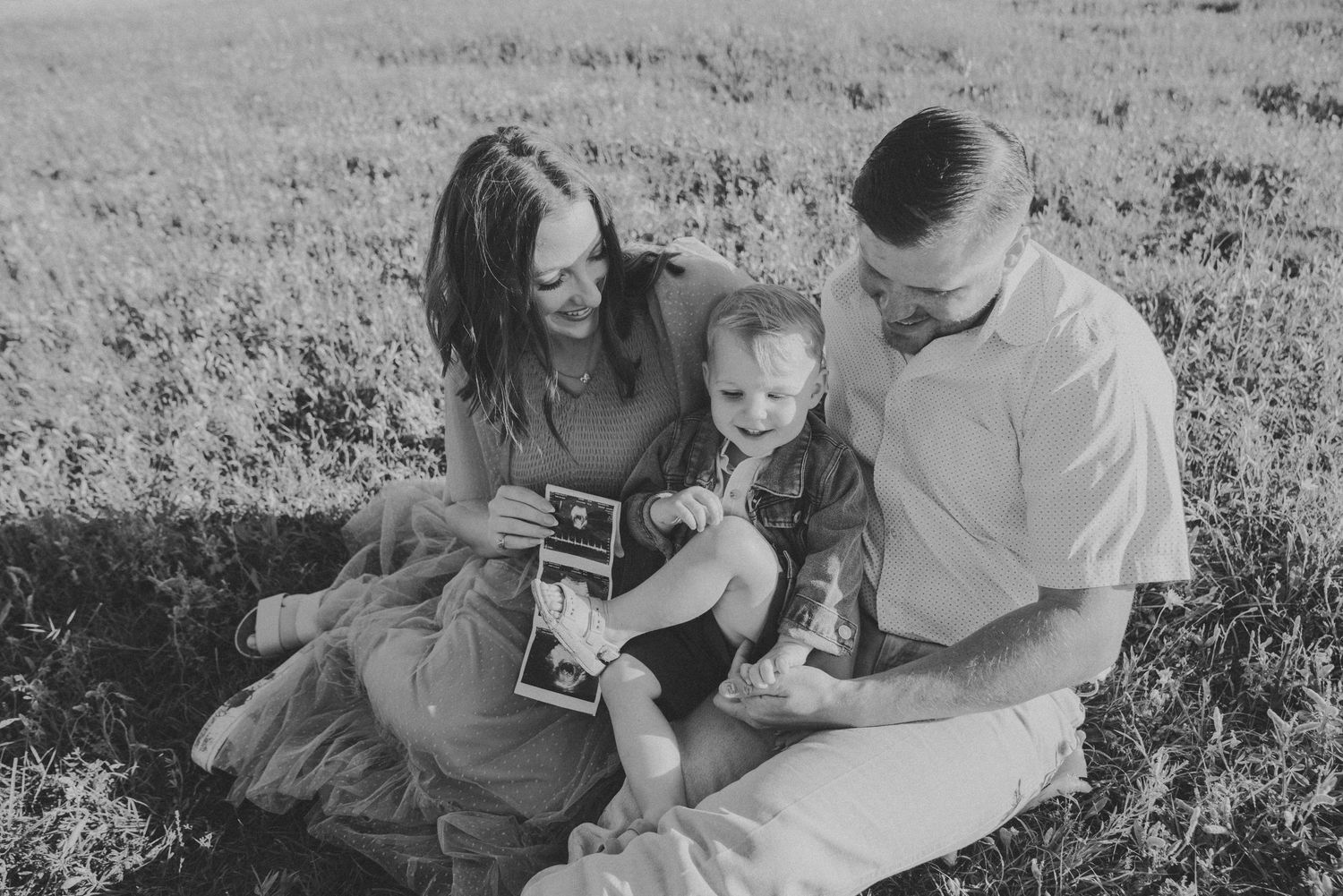 Black and white photo of a couple sitting in a field with their toddler, looking at ultrasound photos together.