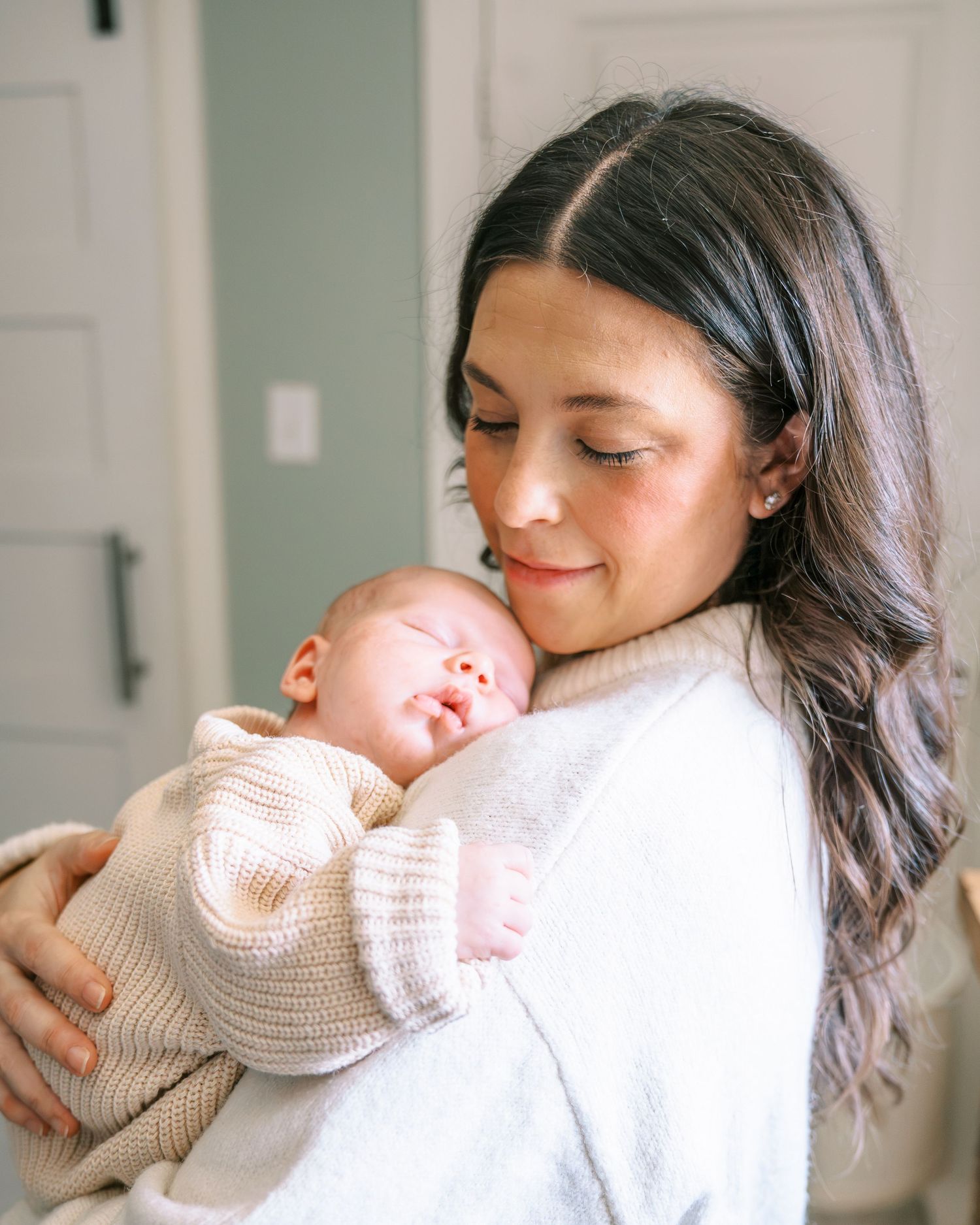 A young mother with long dark hair smiles warmly while cradling her sleeping newborn baby dressed in a beige knit outfit.