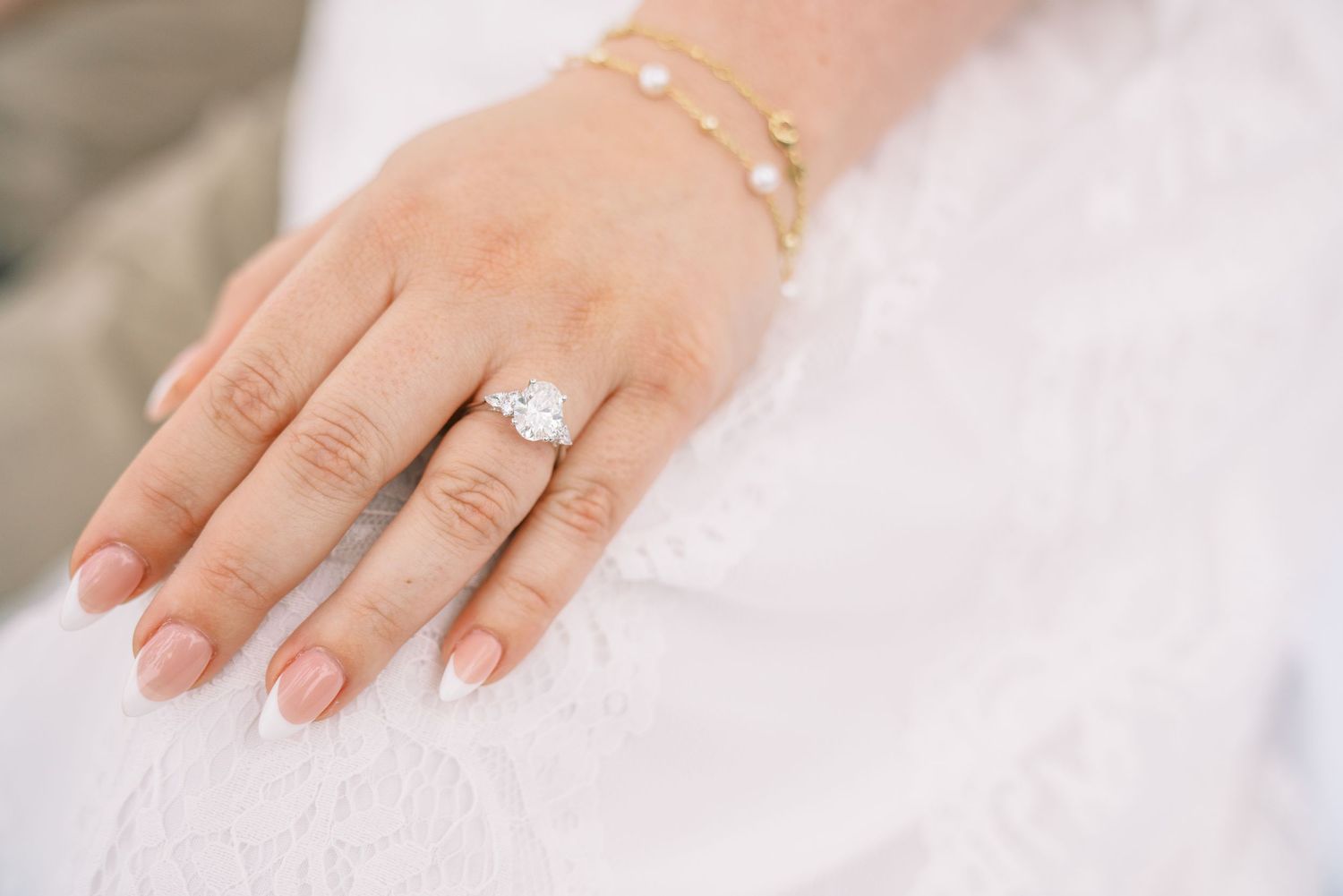 Close-up of a bride's hand wearing a diamond halo engagement ring and gold pearl bracelet on white dress.