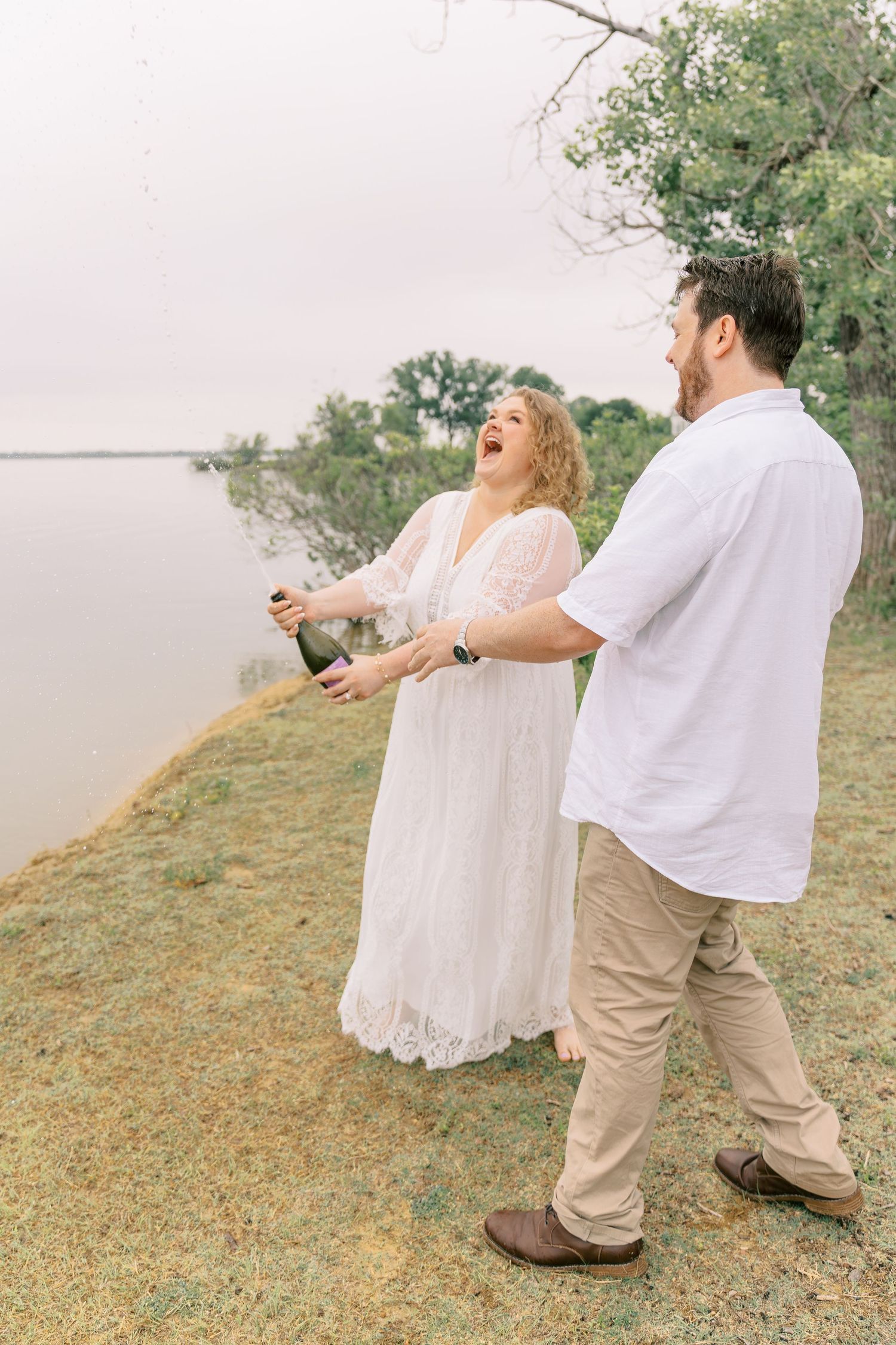 Bride laughing with delight as champagne bottle is opened near a scenic lakeside view.