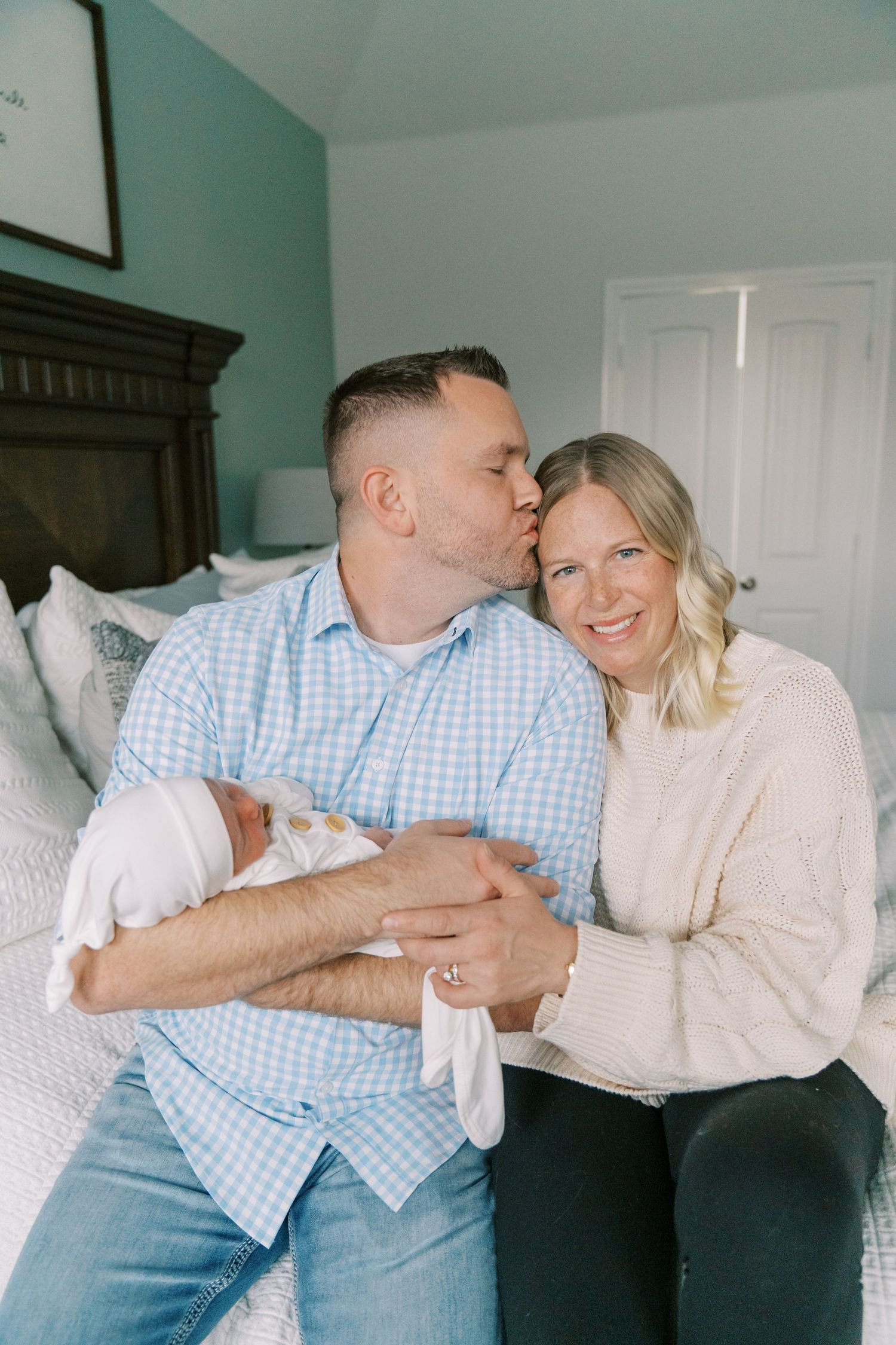 Parents share loving embrace while holding newborn baby in bedroom setting.