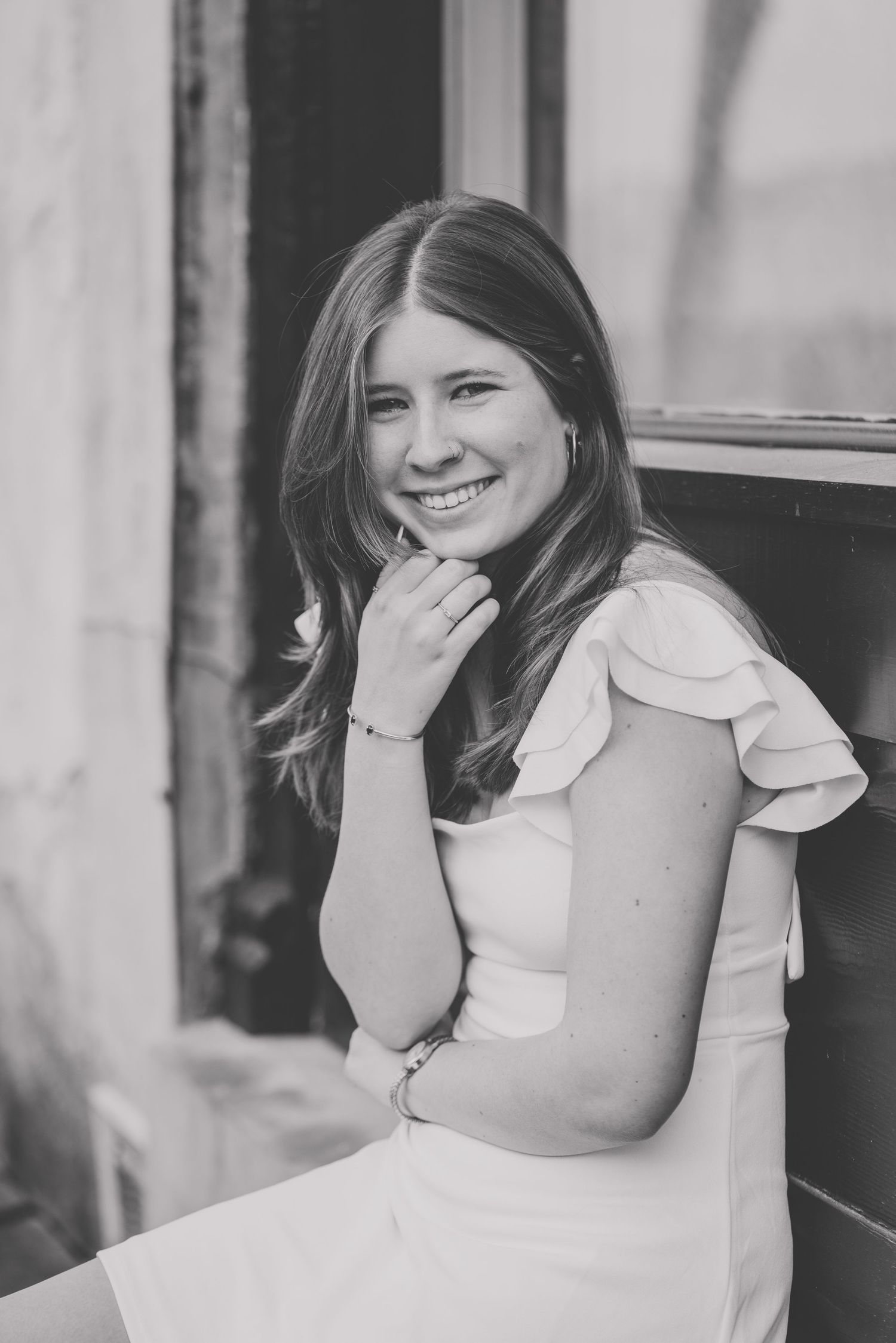 Black and white portrait of a person in a light colored dress with ruffled sleeves smiling while posing against a wall.