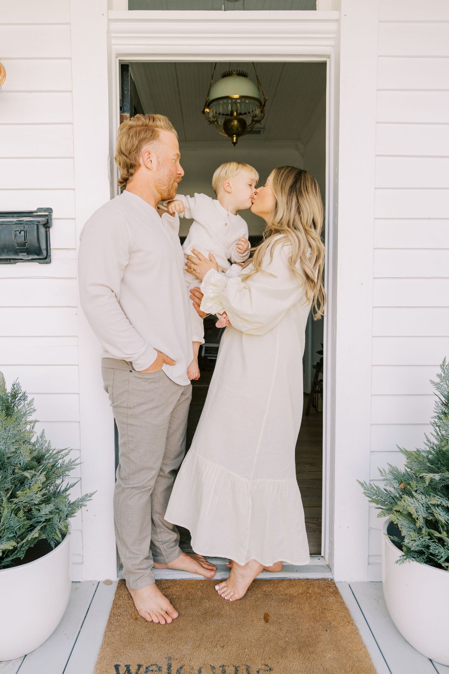 A family shares joyful moments on their white porch with potted plants, dressed in light neutral colors.