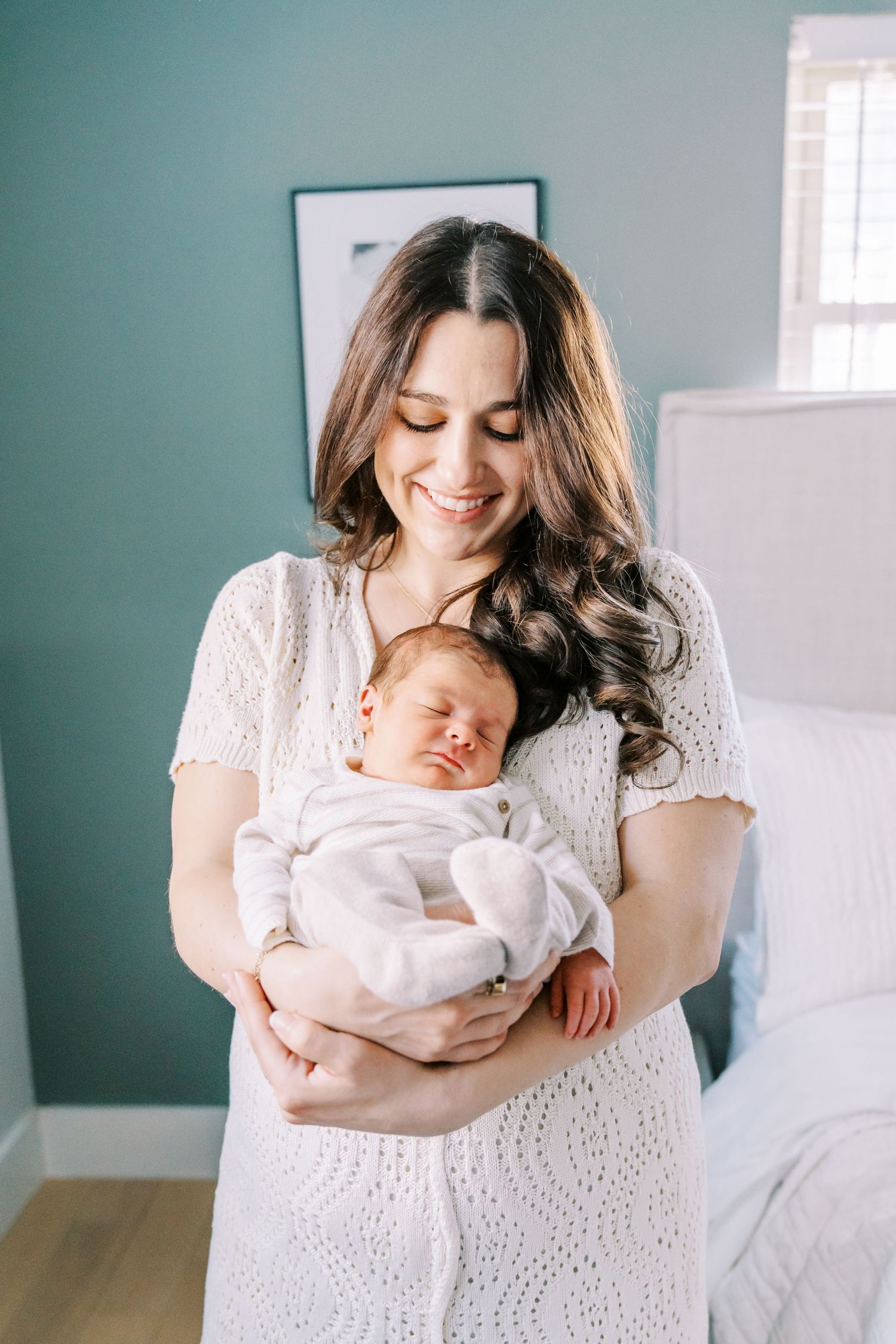 A woman in a white eyelet dress holds and gazes lovingly at a sleeping newborn baby in a bright, airy room.