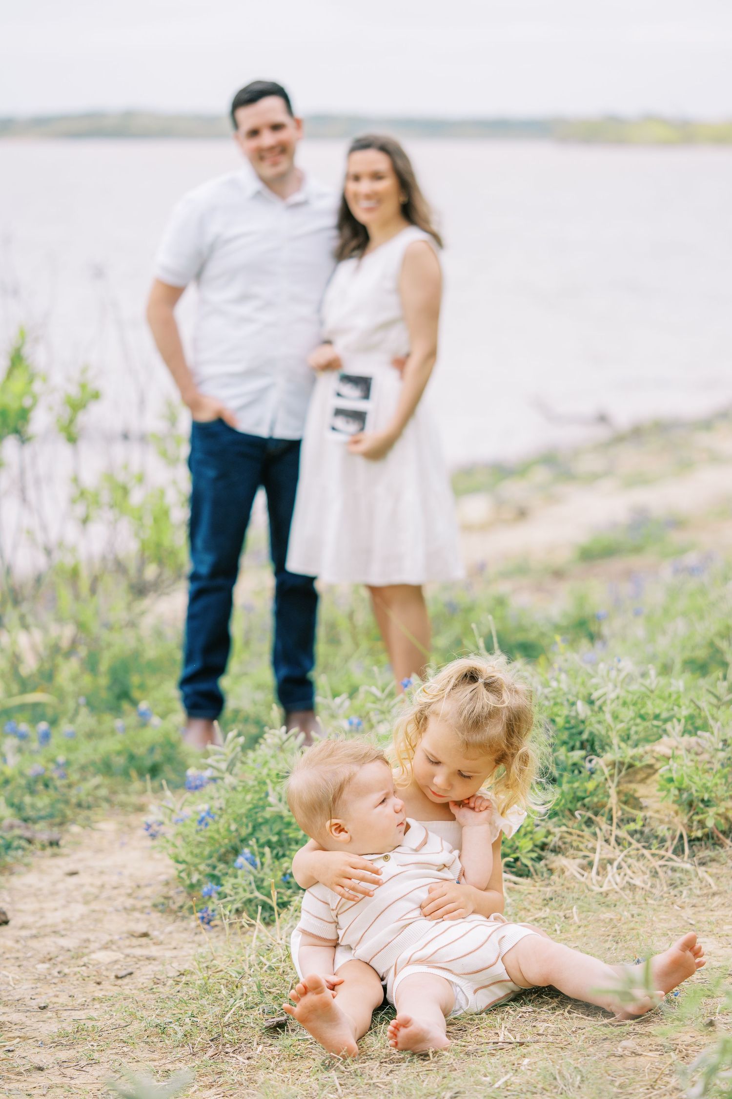 A family maternity photo with two young siblings hugging near a lake, parents holding an ultrasound in the background.