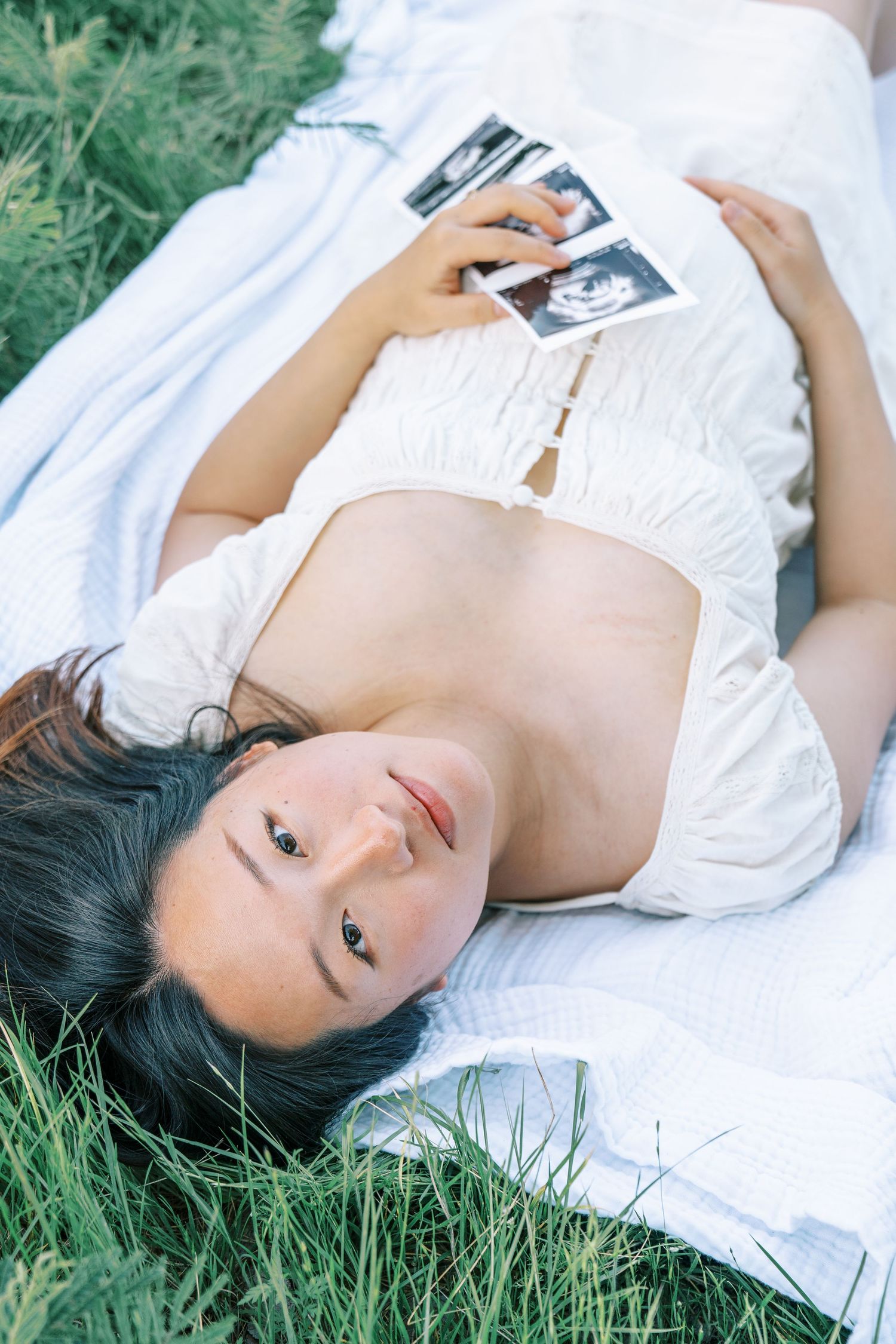 Pregnant woman lying on grass holding ultrasound photos, wearing white crop top, showcasing baby bump outdoors.