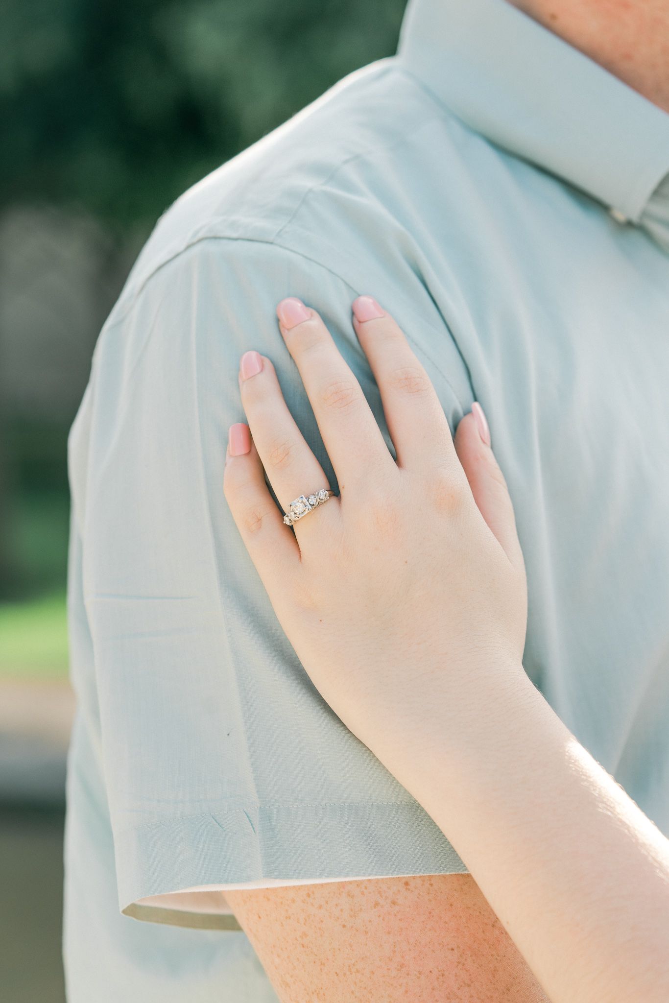 A delicate hand with an engagement ring rests on a light blue shirt sleeve in an outdoor setting.