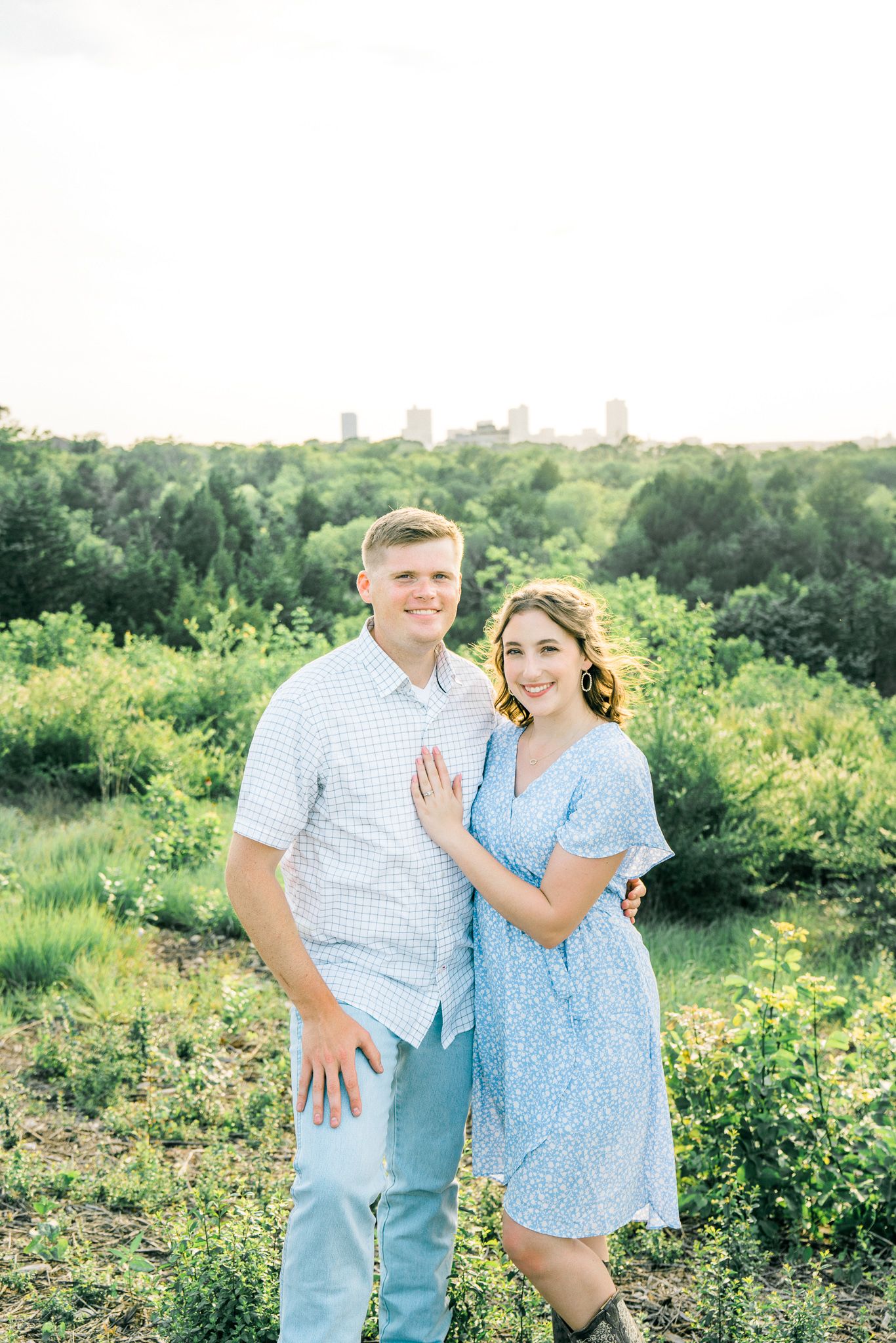A couple stands together on a hilltop overlooking a distant city skyline.