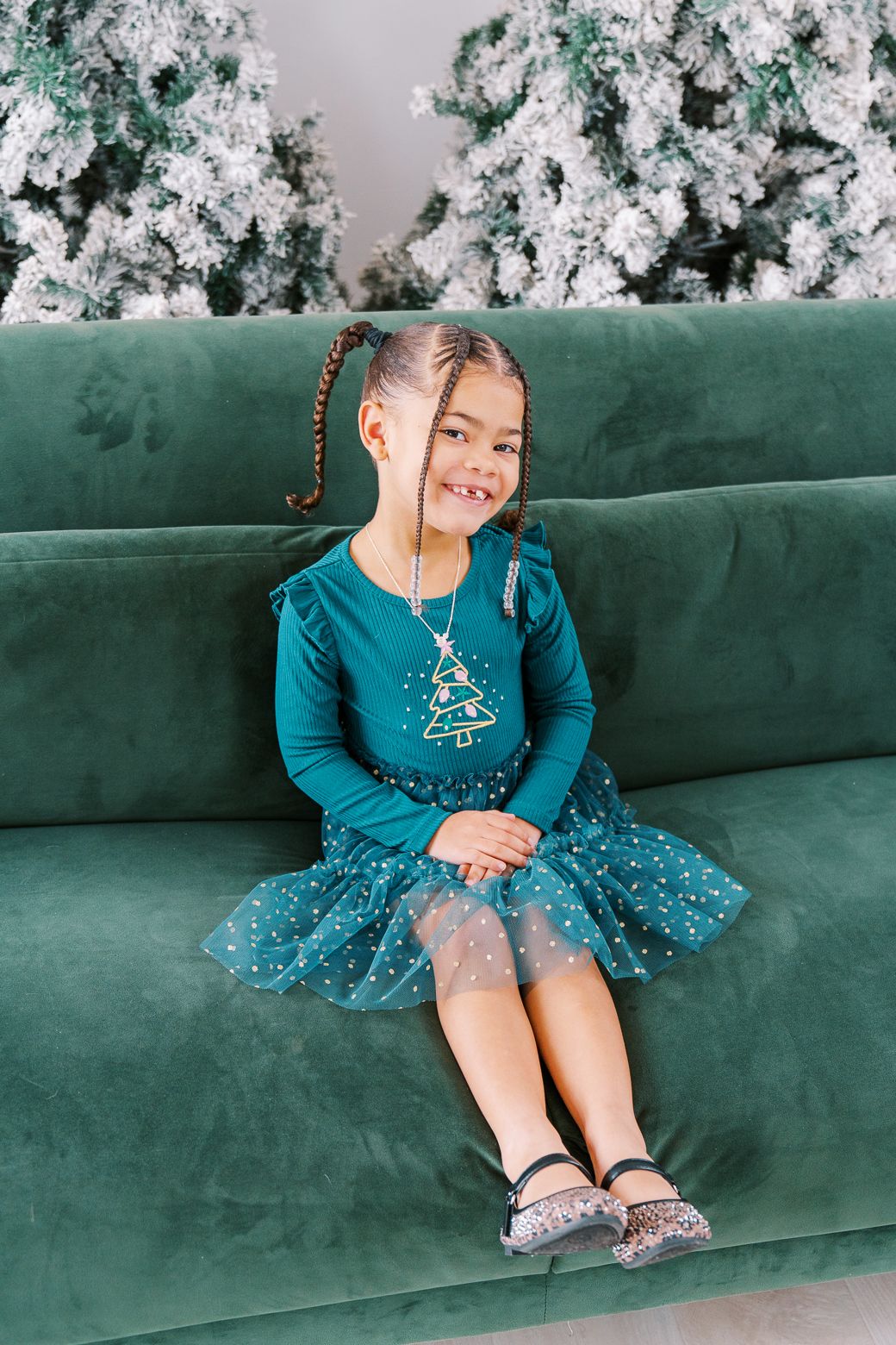 A small child in a sparkly teal dress sits on a green velvet couch with white Christmas decorations in background.