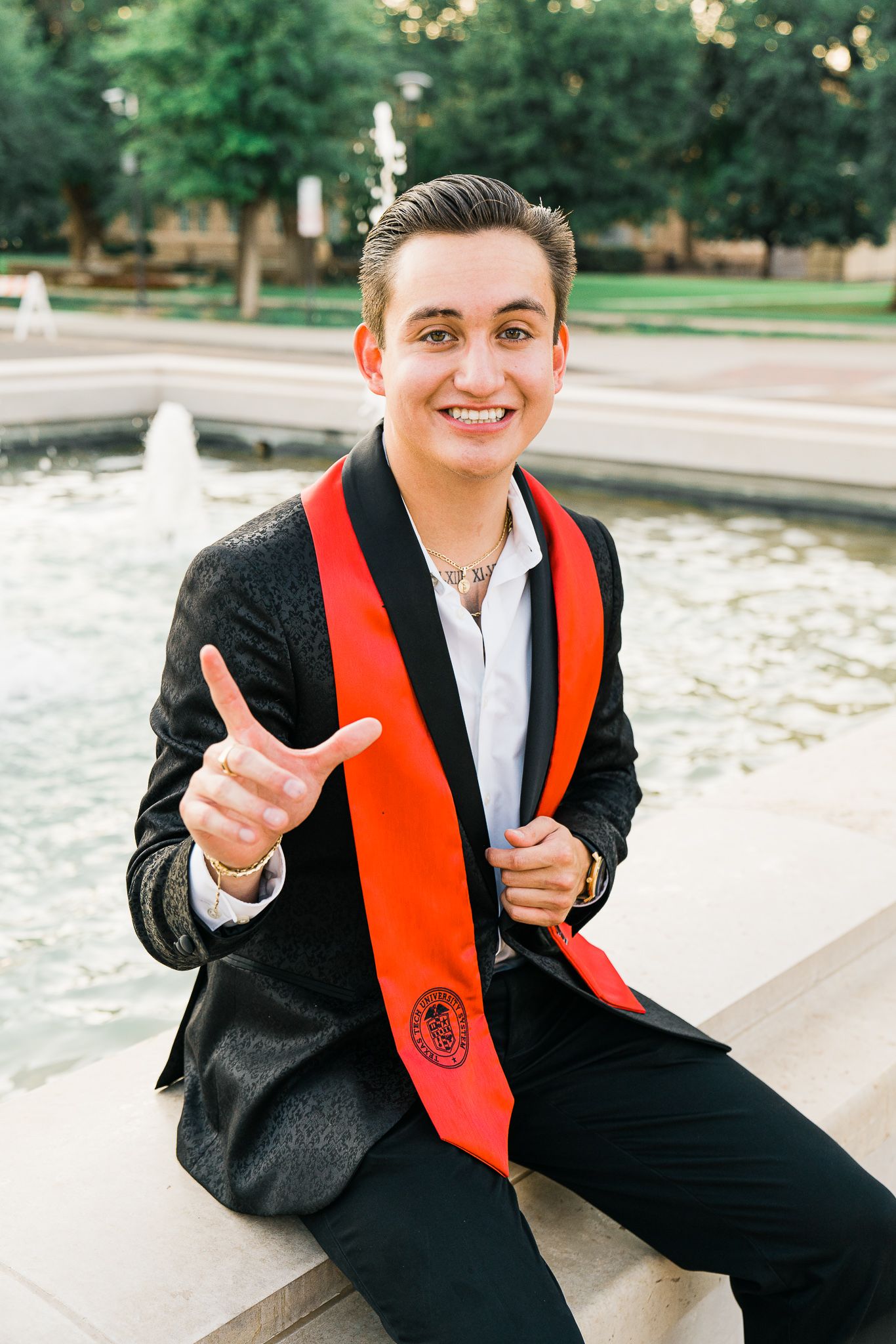 Graduate in black cap and gown with orange stole gives thumbs up while sitting near fountain in outdoor setting.