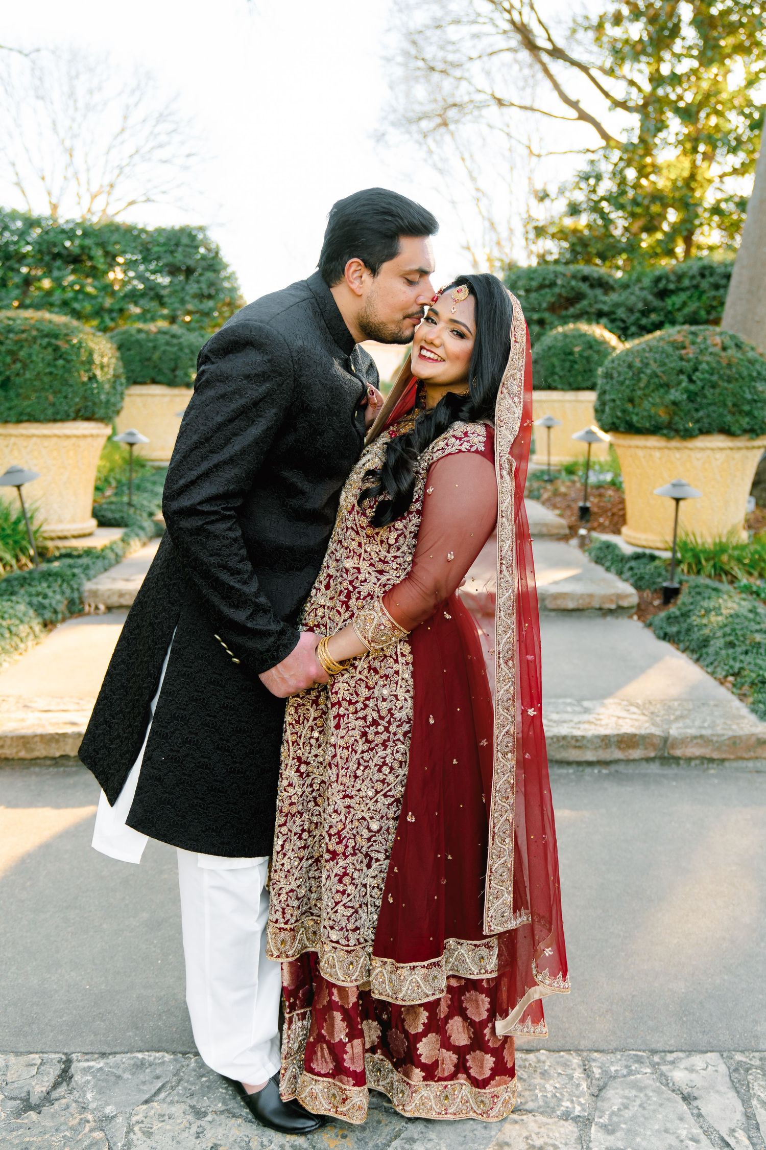 A tender moment between newlyweds in traditional South Asian attire standing in a garden pathway.