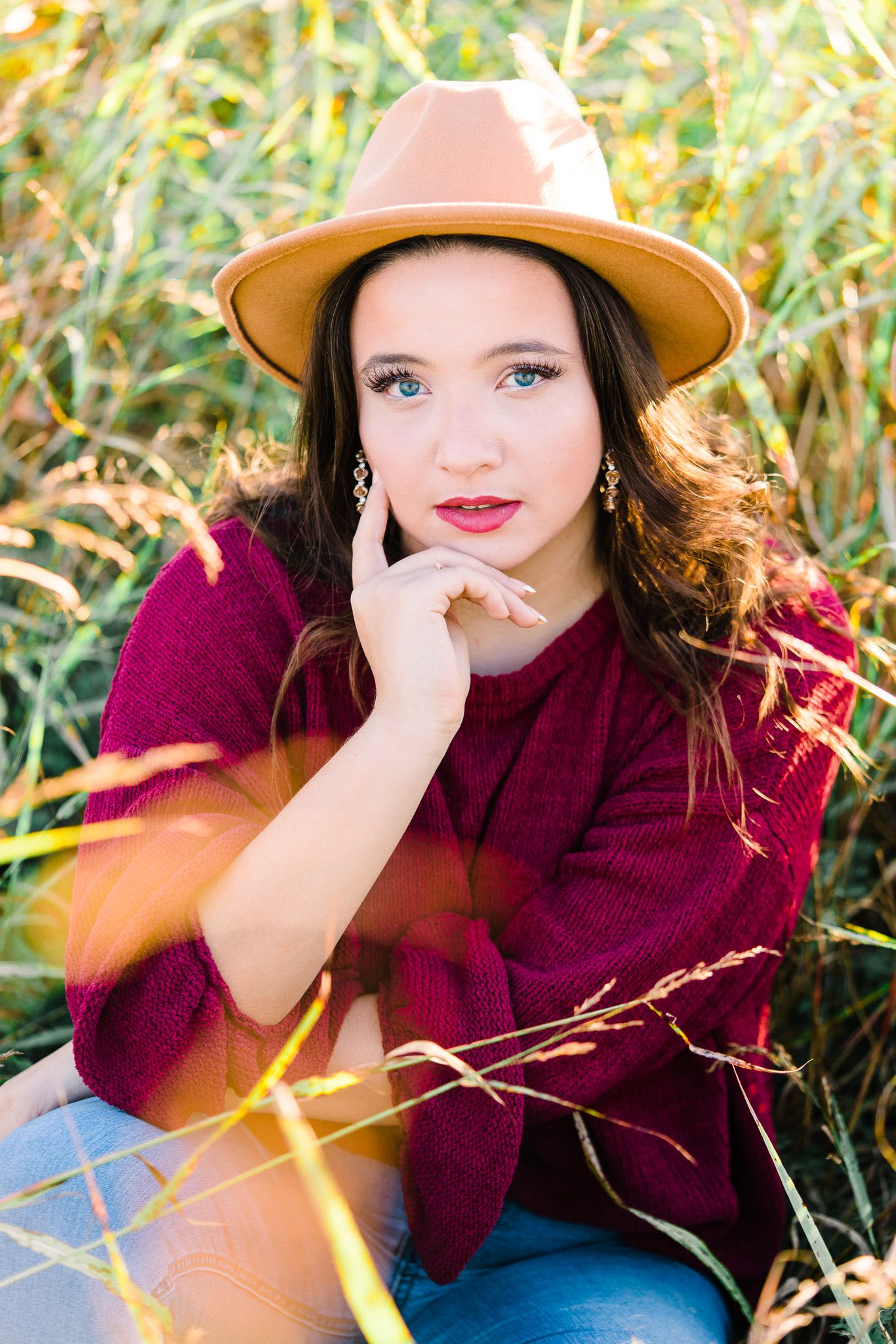 Autumn portrait in burgundy sweater and tan hat sitting in golden grass during sunset.