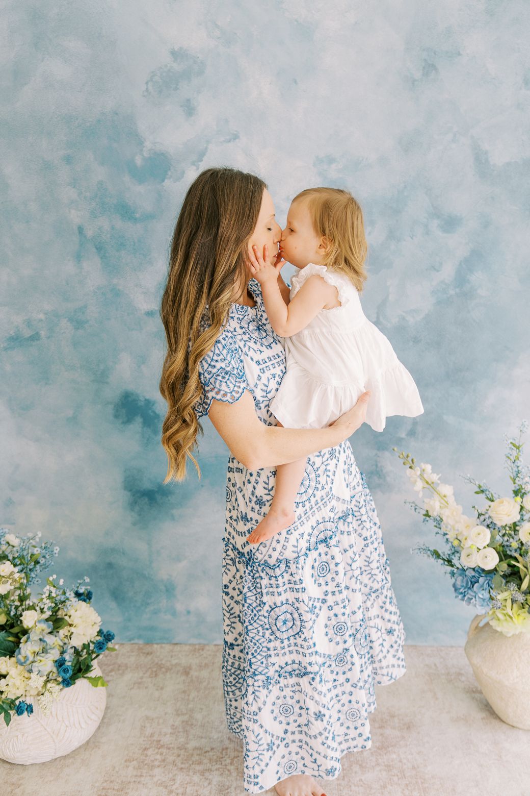 Mother and child in matching white and blue floral dresses embrace in front of a textured blue backdrop with white flowers.