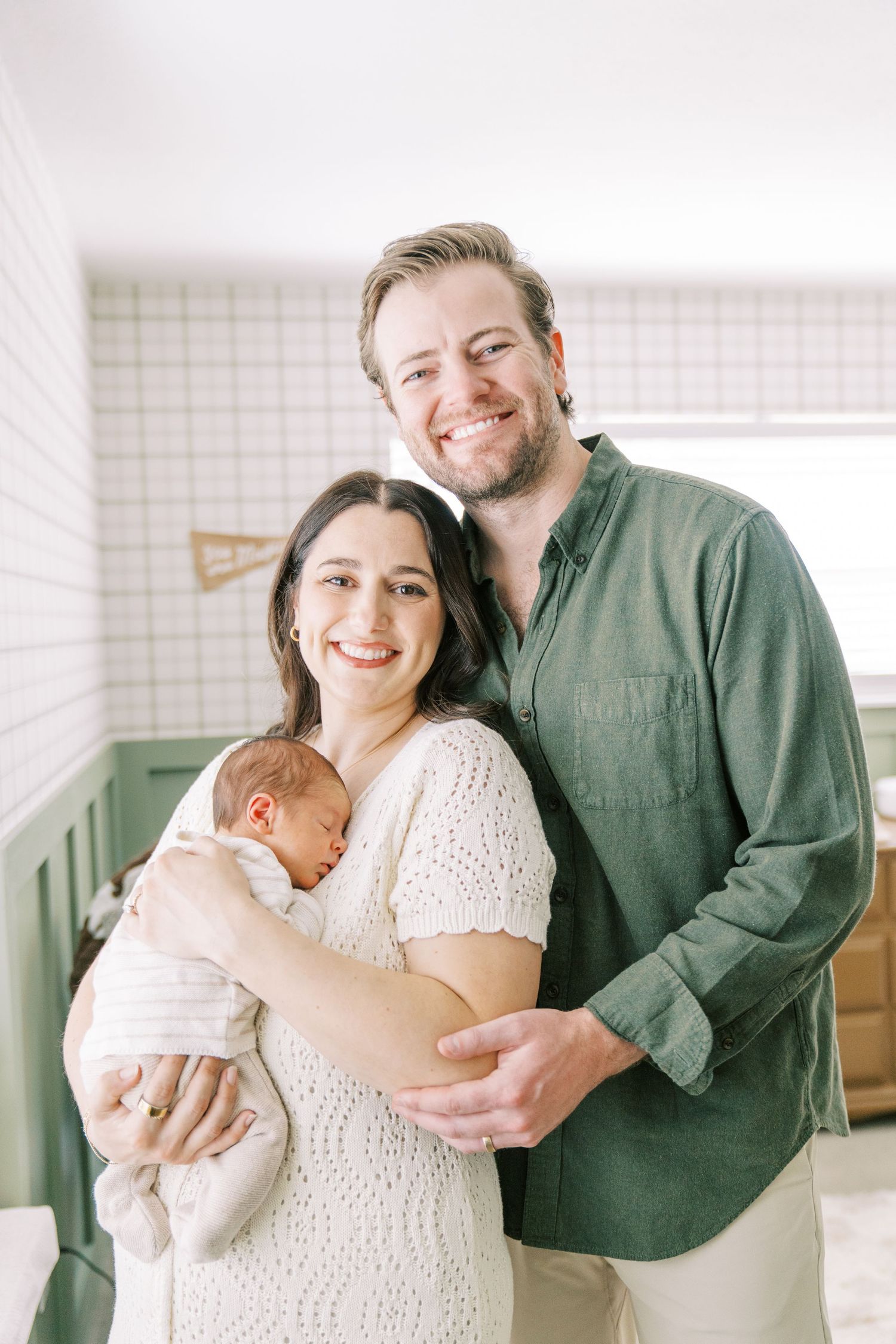 A couple stands together in a bright room while holding their sleeping newborn baby against a white and green background.