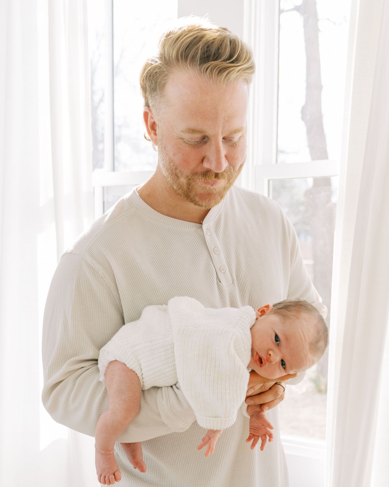 Parent and baby in white clothing share playful interactions against white curtains.