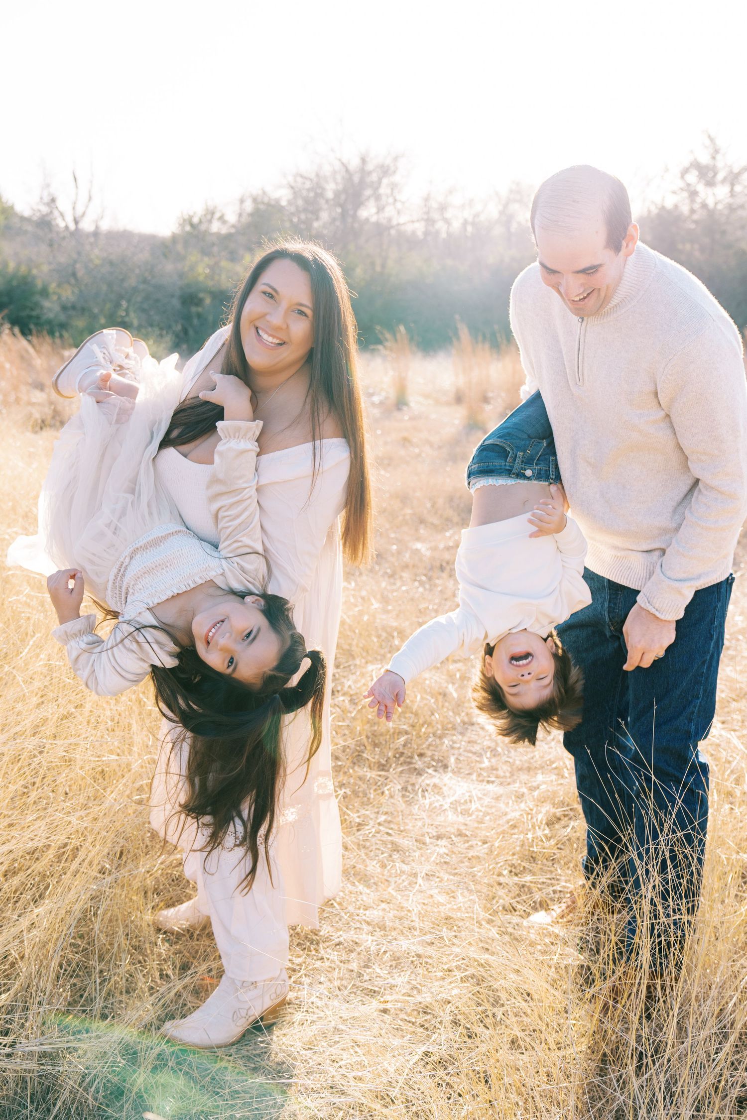 Playful family moment captured in sunlit field wearing coordinated white outfits.