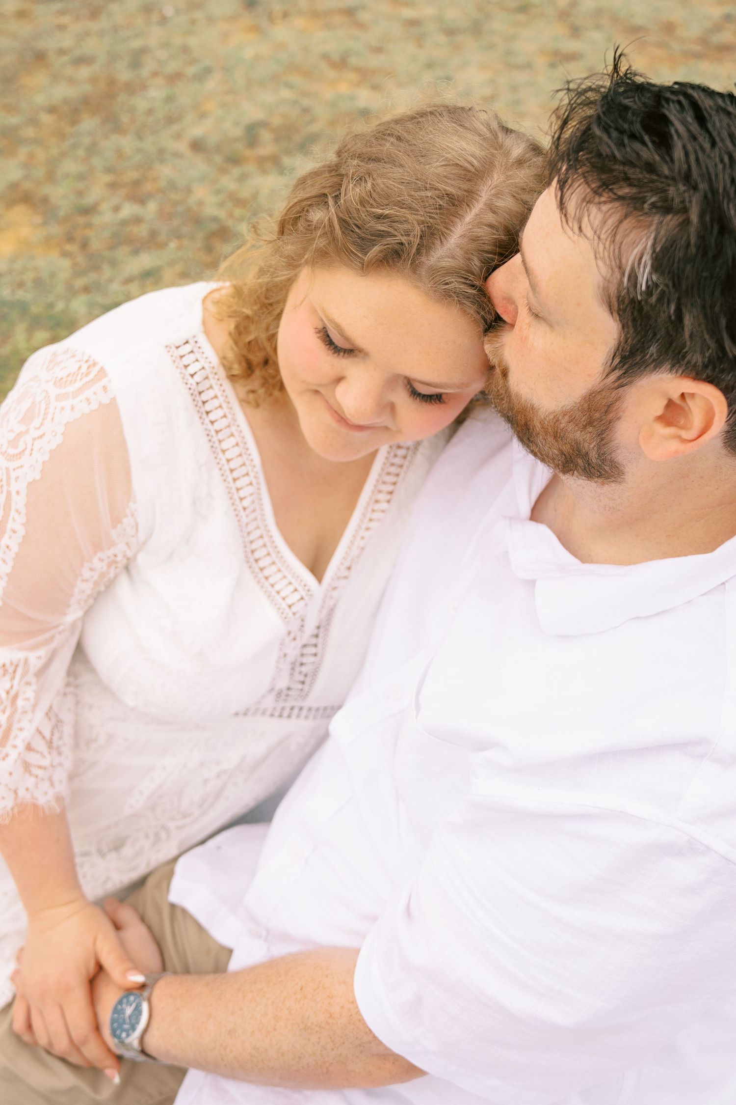 A bearded man tenderly kisses a woman's temple as they sit together during an engagement photo session.