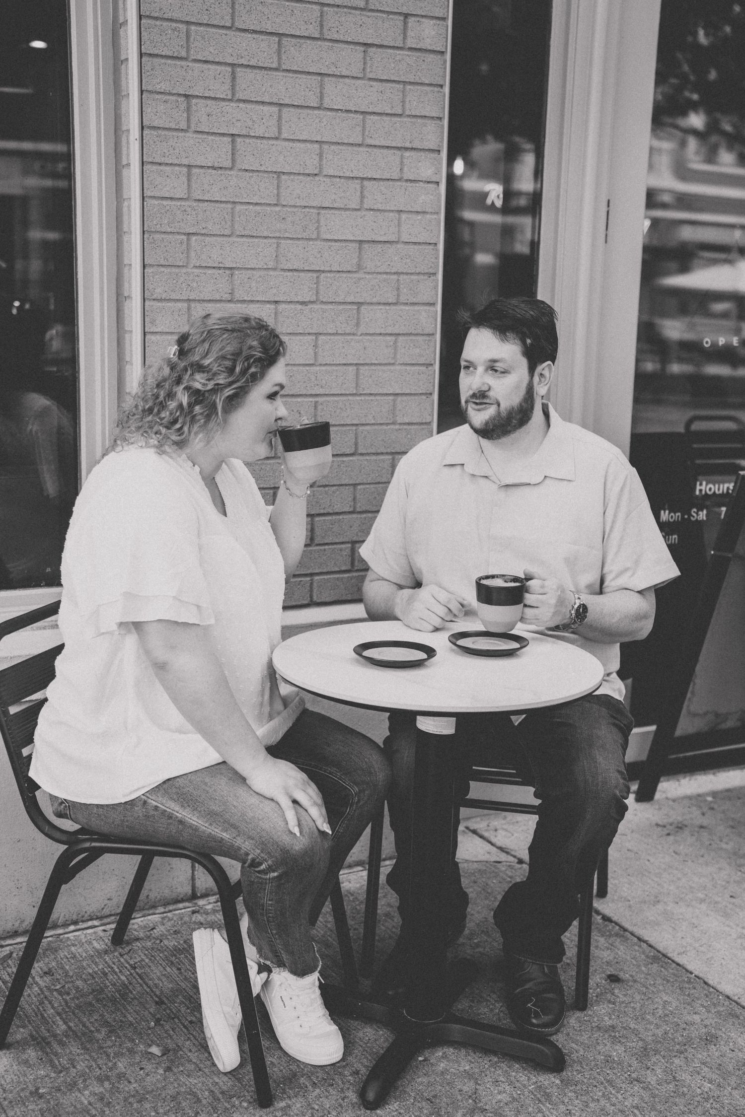Black and white photo of engaged couple enjoying coffee together at outdoor cafe table.