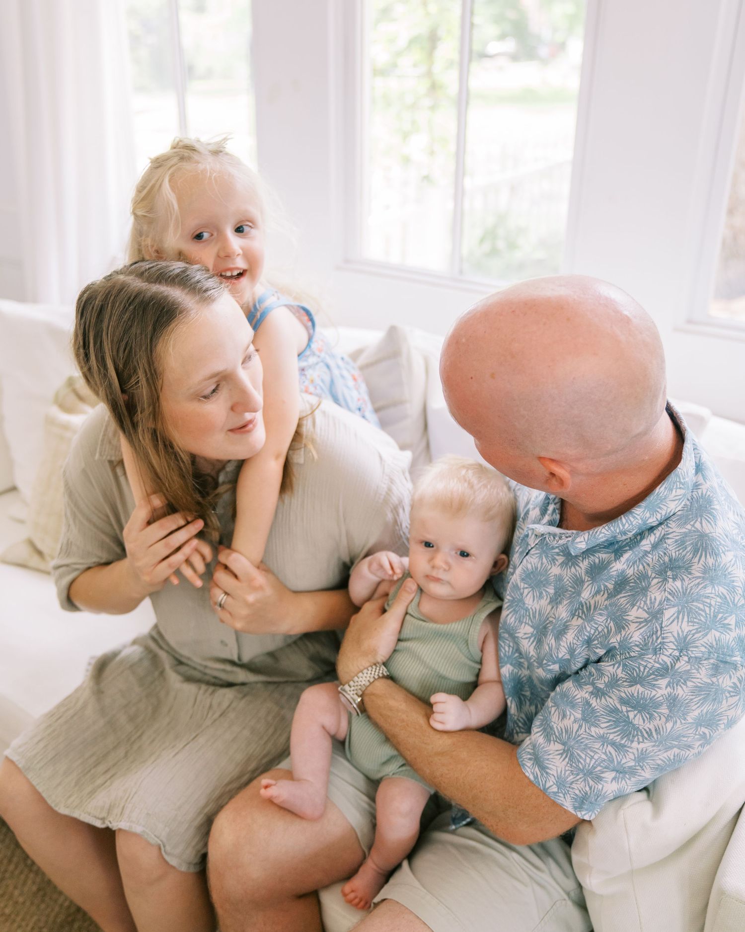 A happy family of four sits together on a white couch, with parents smiling at their toddler and baby in a bright room.