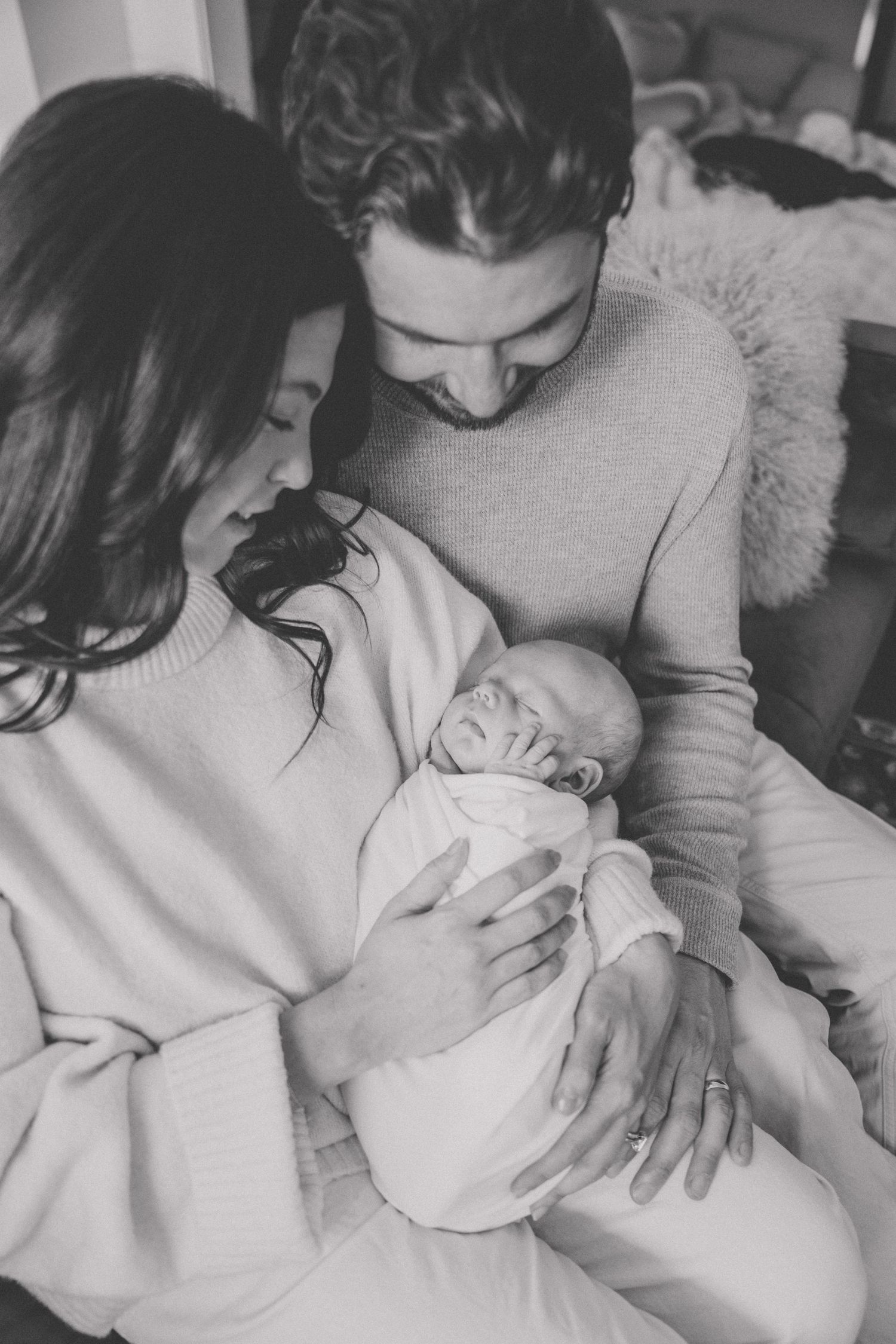 Black and white photo of new parents lovingly gazing at their swaddled newborn baby.