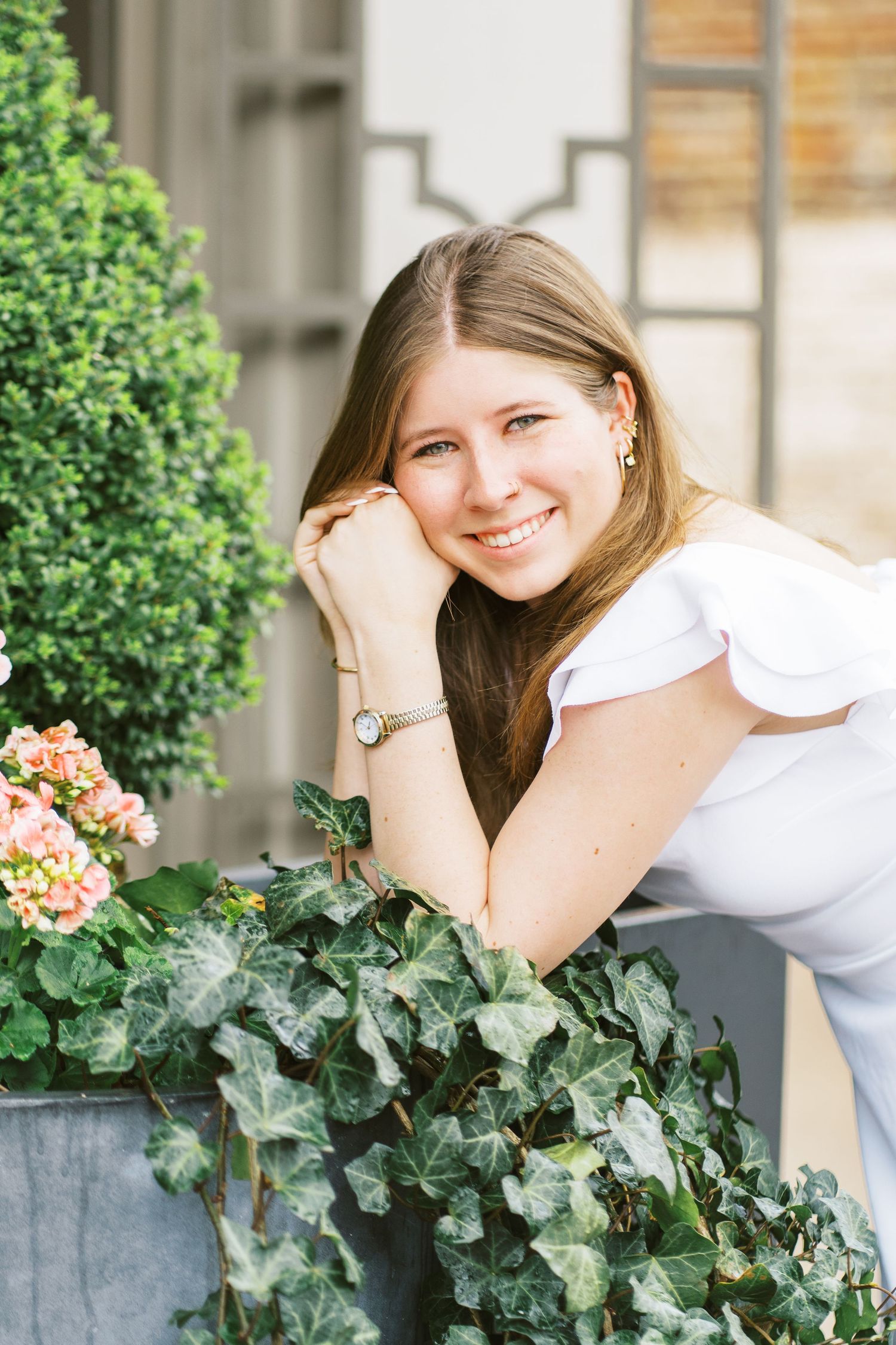 A person in a white top smiles while leaning on a planter with ivy and flowers outside a building.
