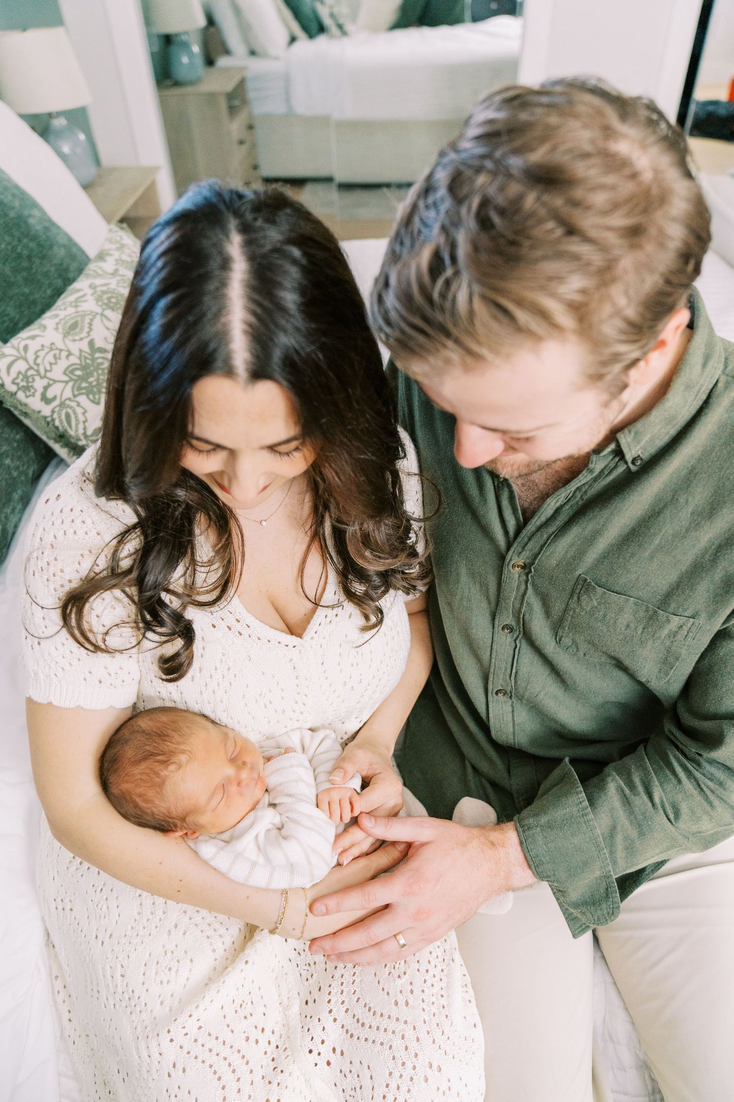 A couple in casual attire lovingly holds their sleeping newborn baby while sitting together indoors.