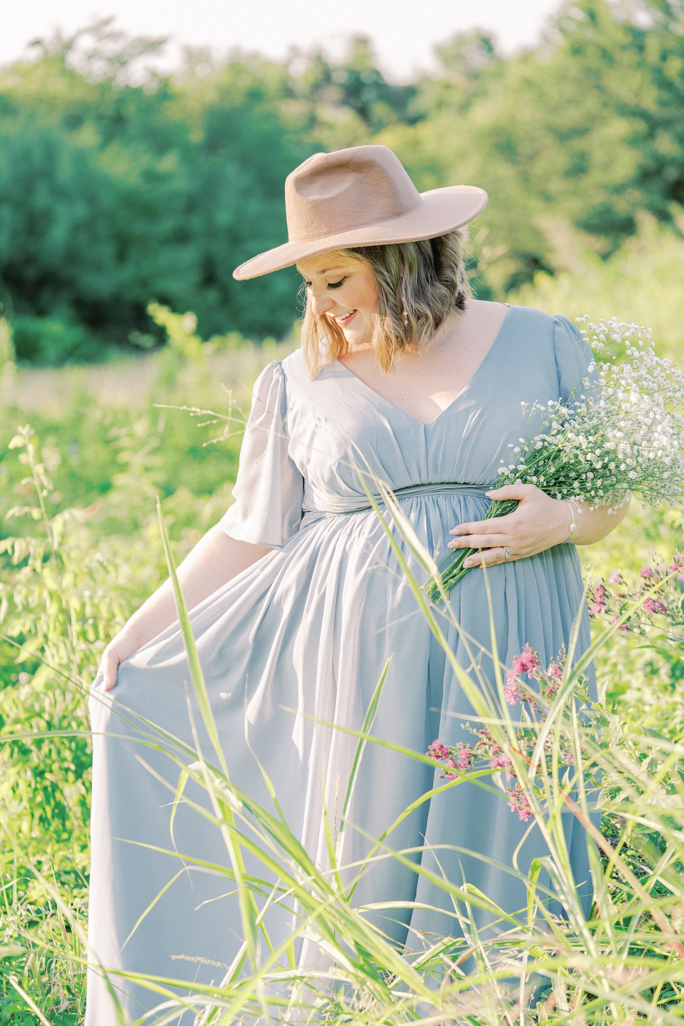 A woman in a flowing blue dress and wide-brimmed hat gathers wildflowers in a sunlit meadow.