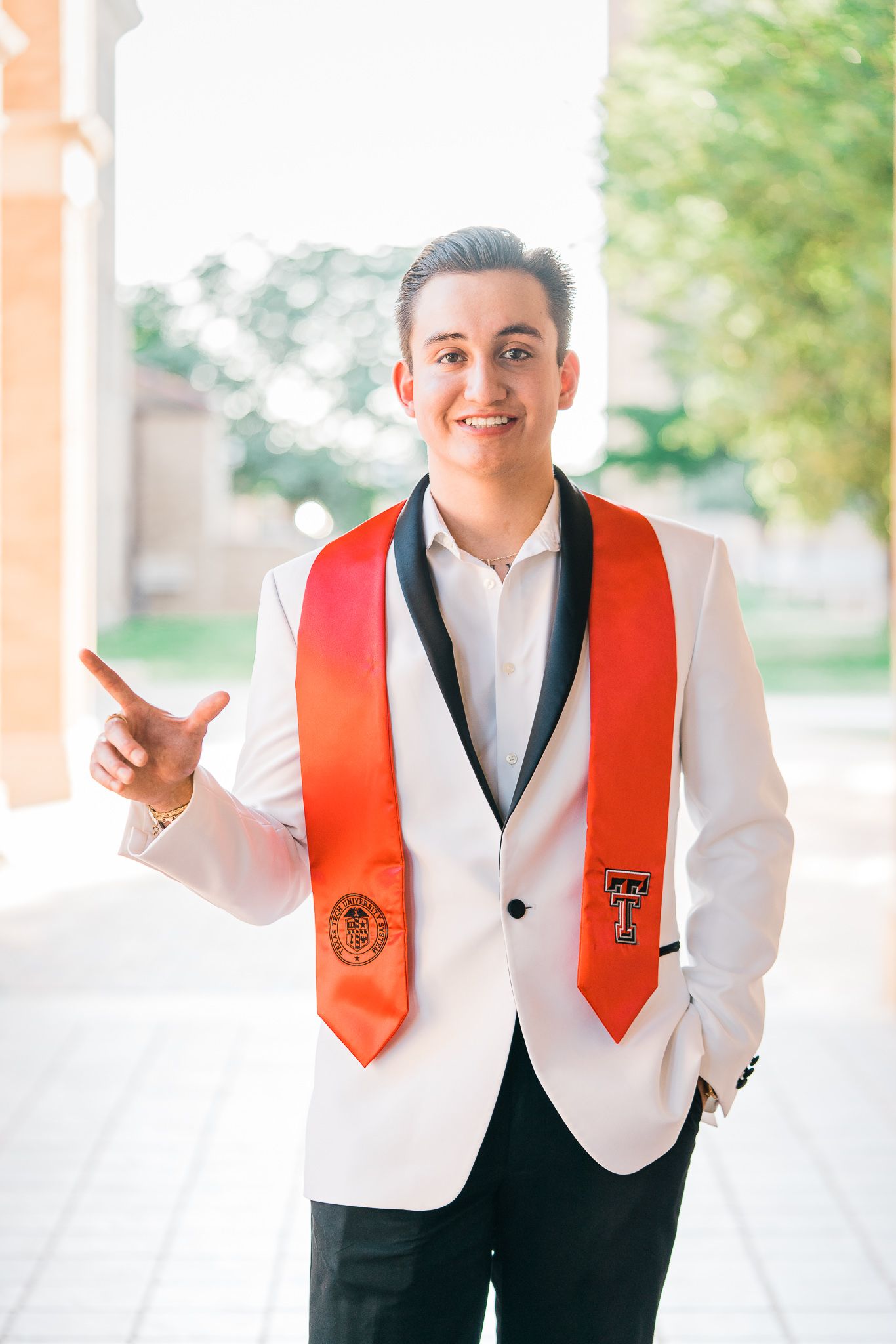 Graduate in white jacket and red stole gestures playfully during graduation photo session.