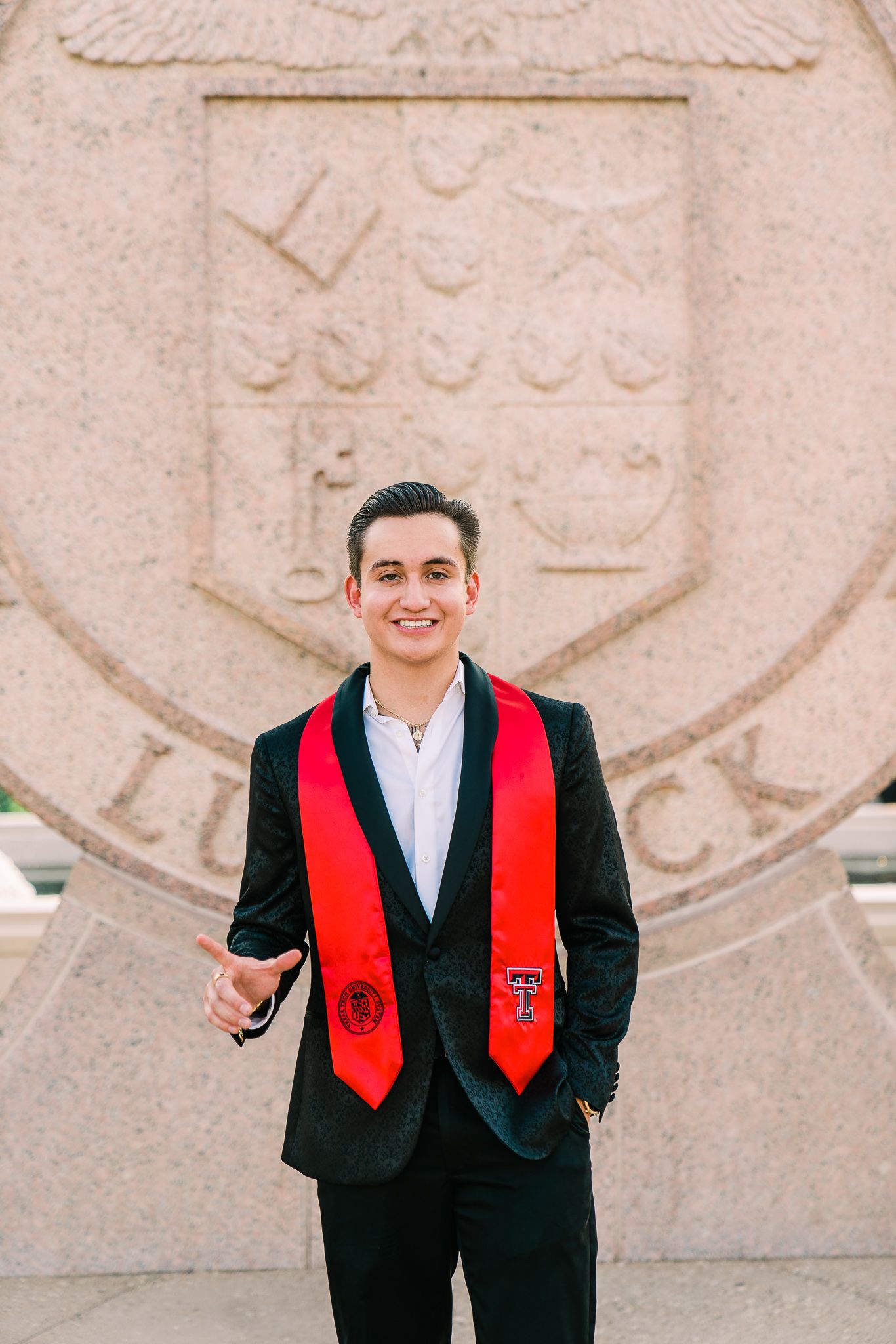 A graduate in formal attire with a red stole stands in front of a stone university crest.