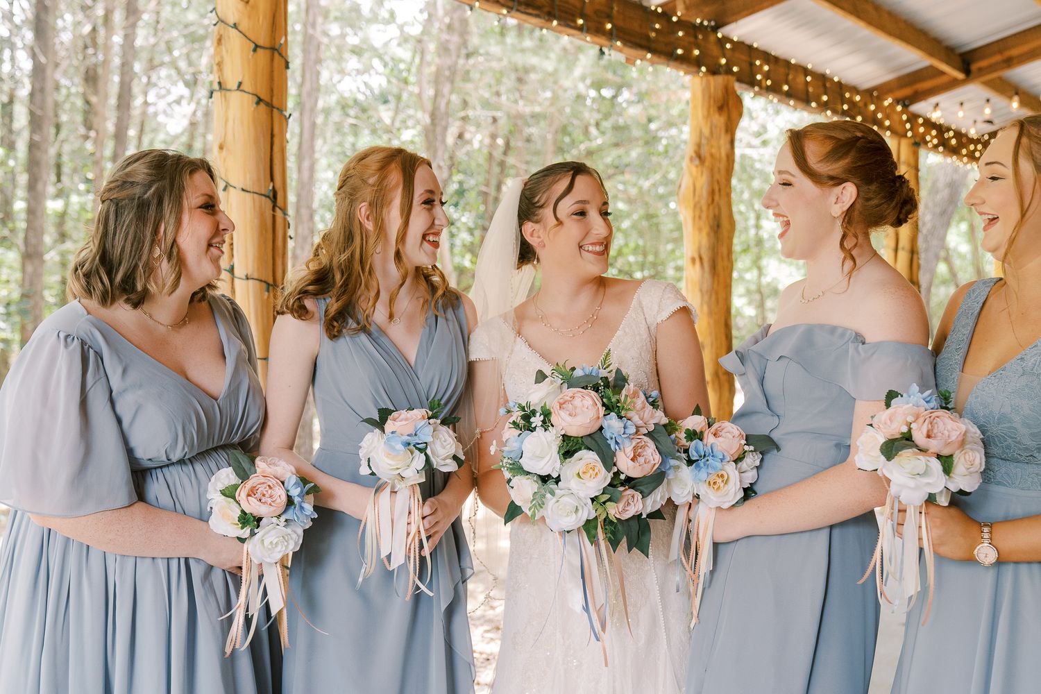 Bridesmaids in matching dusty blue dresses hold pastel bouquets while standing in a row against a rustic wooden backdrop.