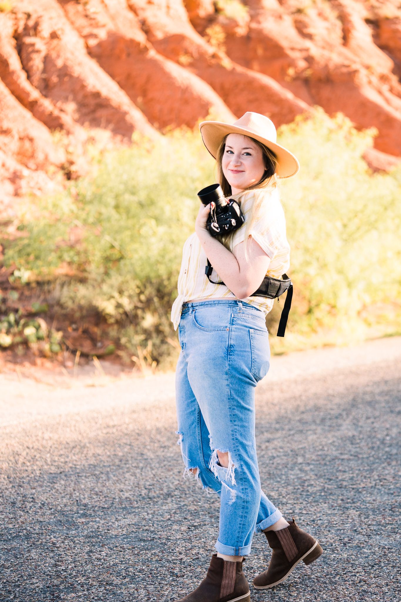 Person in casual western attire with straw hat and boots holding a black dog against red rock formations outdoors.
