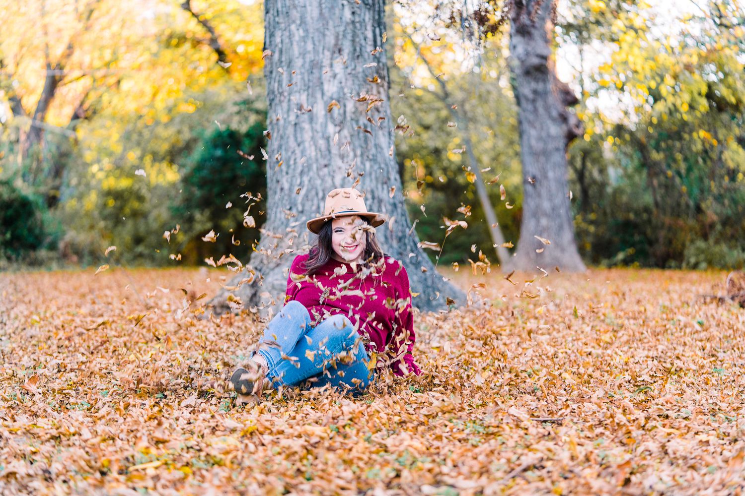 A playful autumn scene with someone laying in vibrant orange fallen leaves beneath tall trees in a park.
